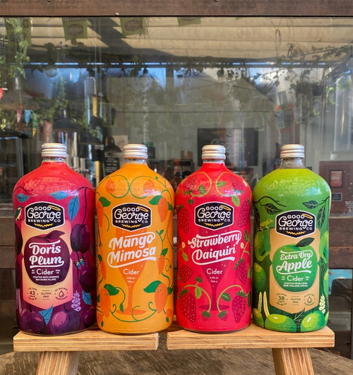 Four bottles of Good George Brewing Co. cider on a wooden stand with a blurred indoor background.