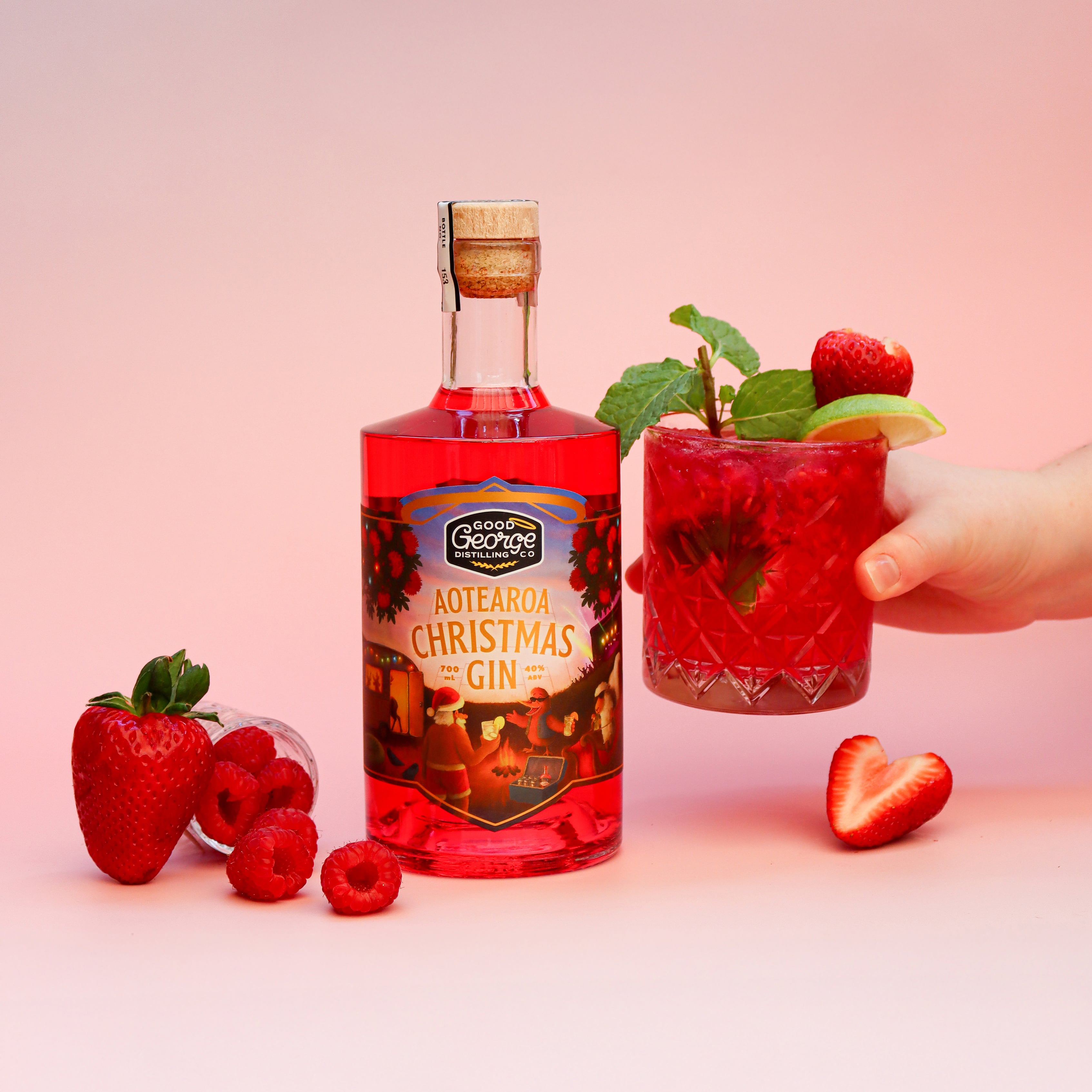 Bottle of Aotearoa Christmas Gin with a glass of red cocktail garnished with strawberries and raspberries on a pink background.