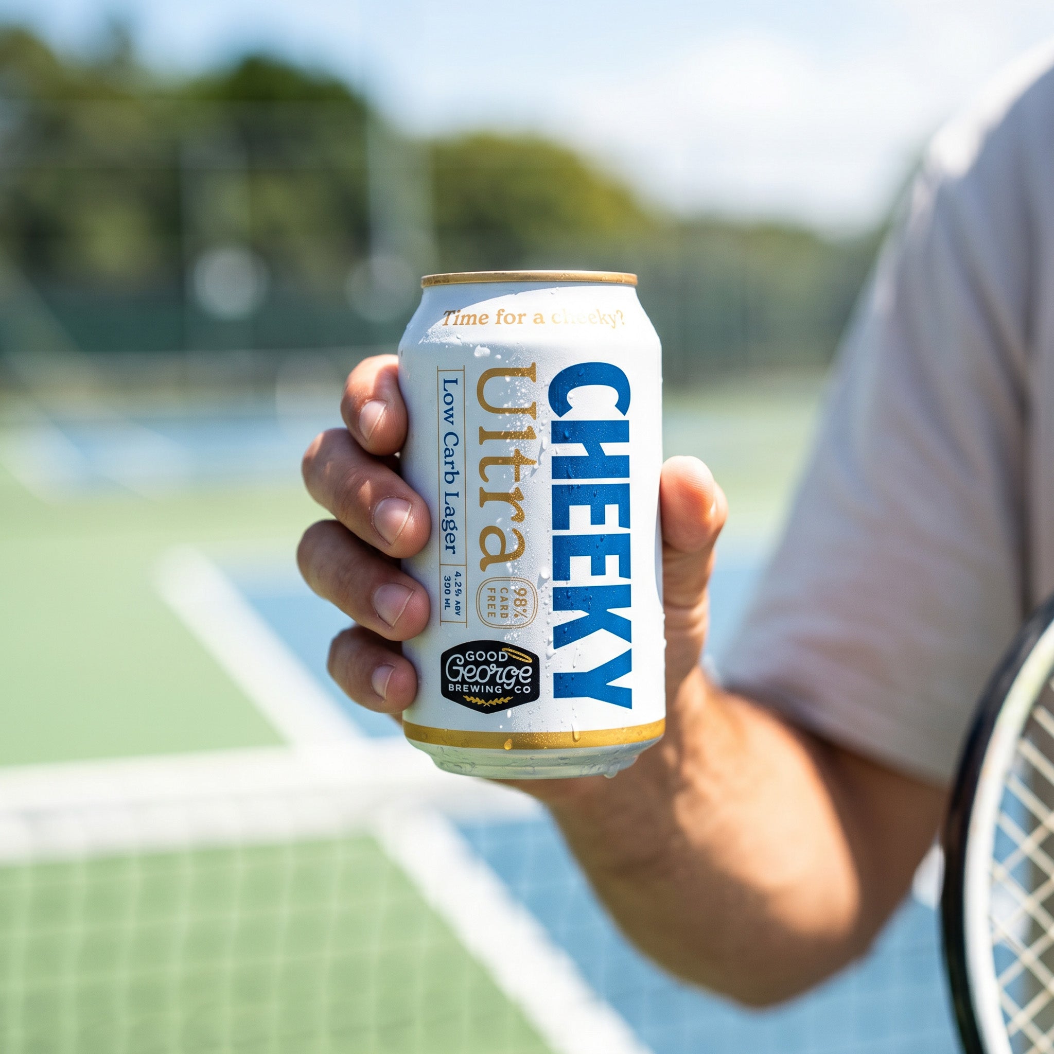 A person holding a can of Cheeky Ultra low carb lager next to a tennis racket on a tennis court.