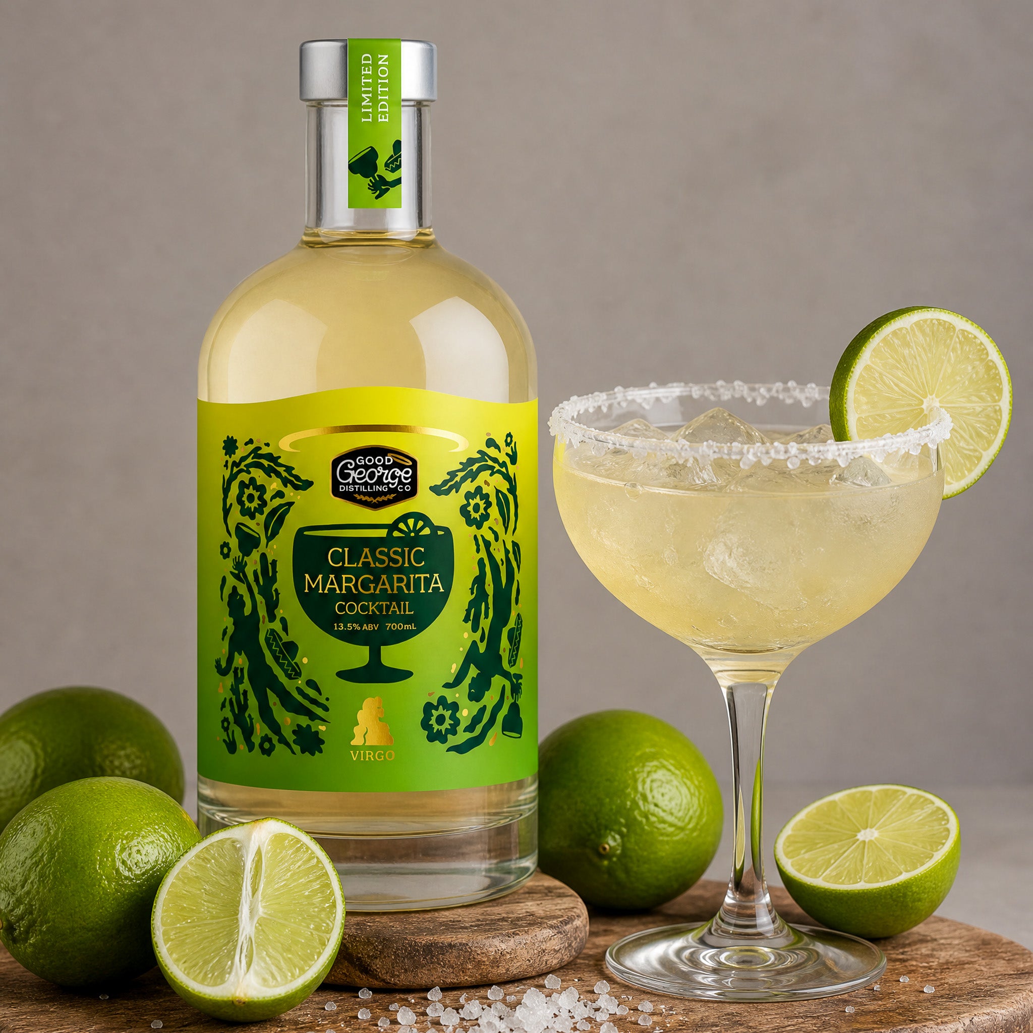 A bottle of Good George Classic Margarita cocktail spirit stands beside a margarita glass with a lime wheel garnish, surrounded by whole and sliced limes on a wooden surface against a neutral gray background.