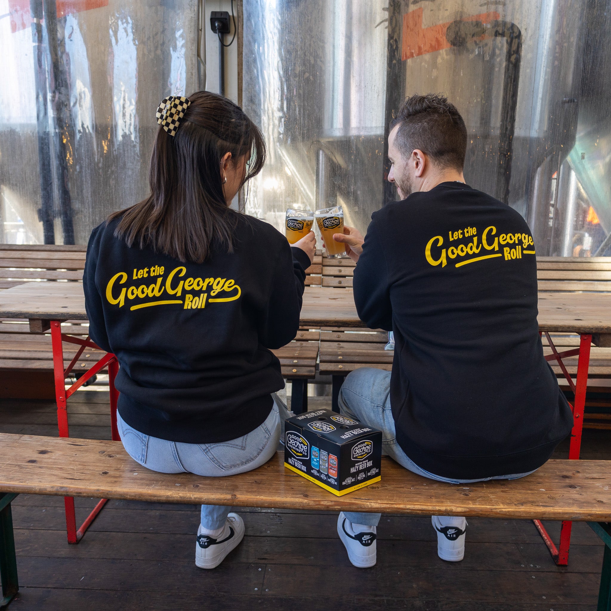 Two individuals sitting on a bench wearing black crew neck sweatshirts with text on them, drinking from glasses.