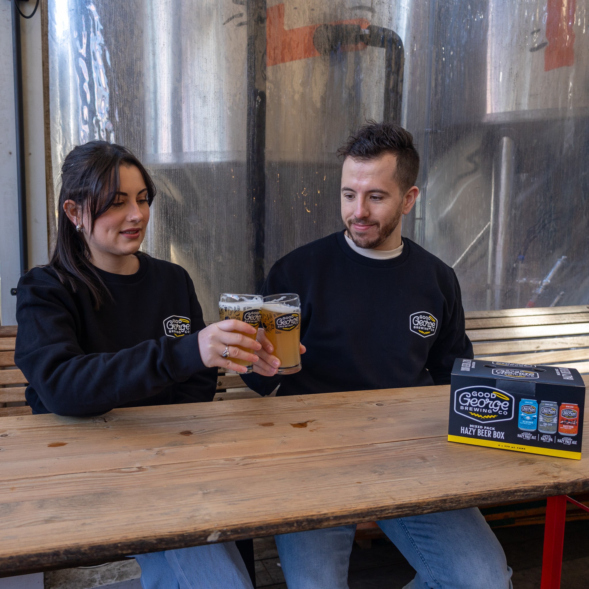 Two individuals sitting on a bench wearing black crew neck sweatshirts with text on them, drinking from glasses.
