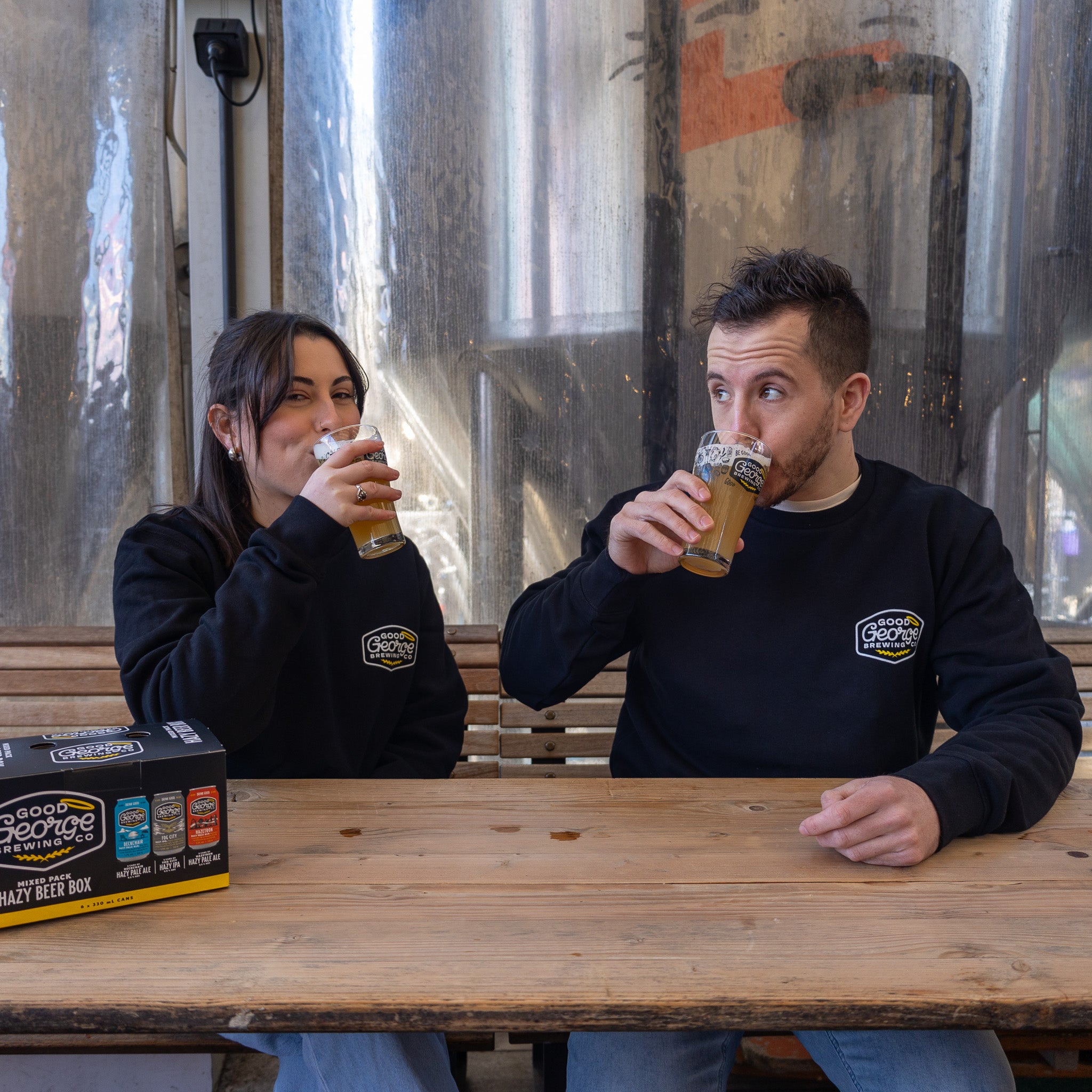 Two individuals sitting on a bench wearing black crew neck sweatshirts with text on them, drinking from glasses.