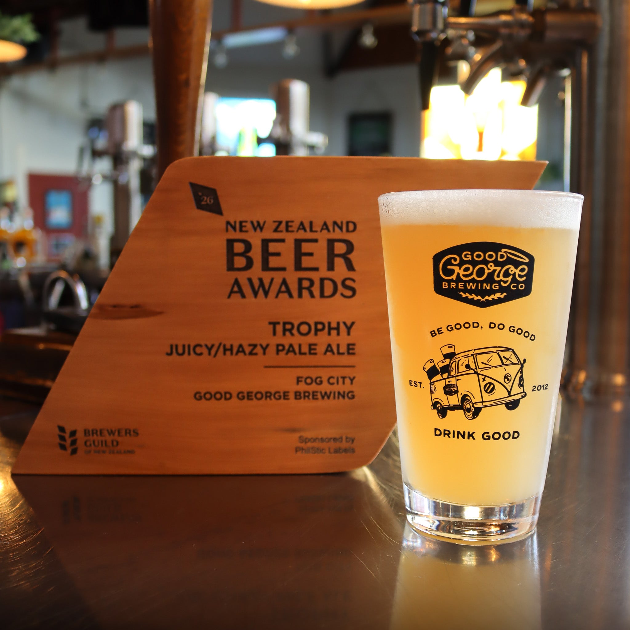 A pint of Hazy pale ale with a white foam head sits on a bar counter beside a wooden New Zealand Beer Awards trophy sign. The pint glass is branded Good George Brewing Co with a cartoon van graphic and the words 'BE GOOD, DO GOOD' and 'DRINK GOOD'.