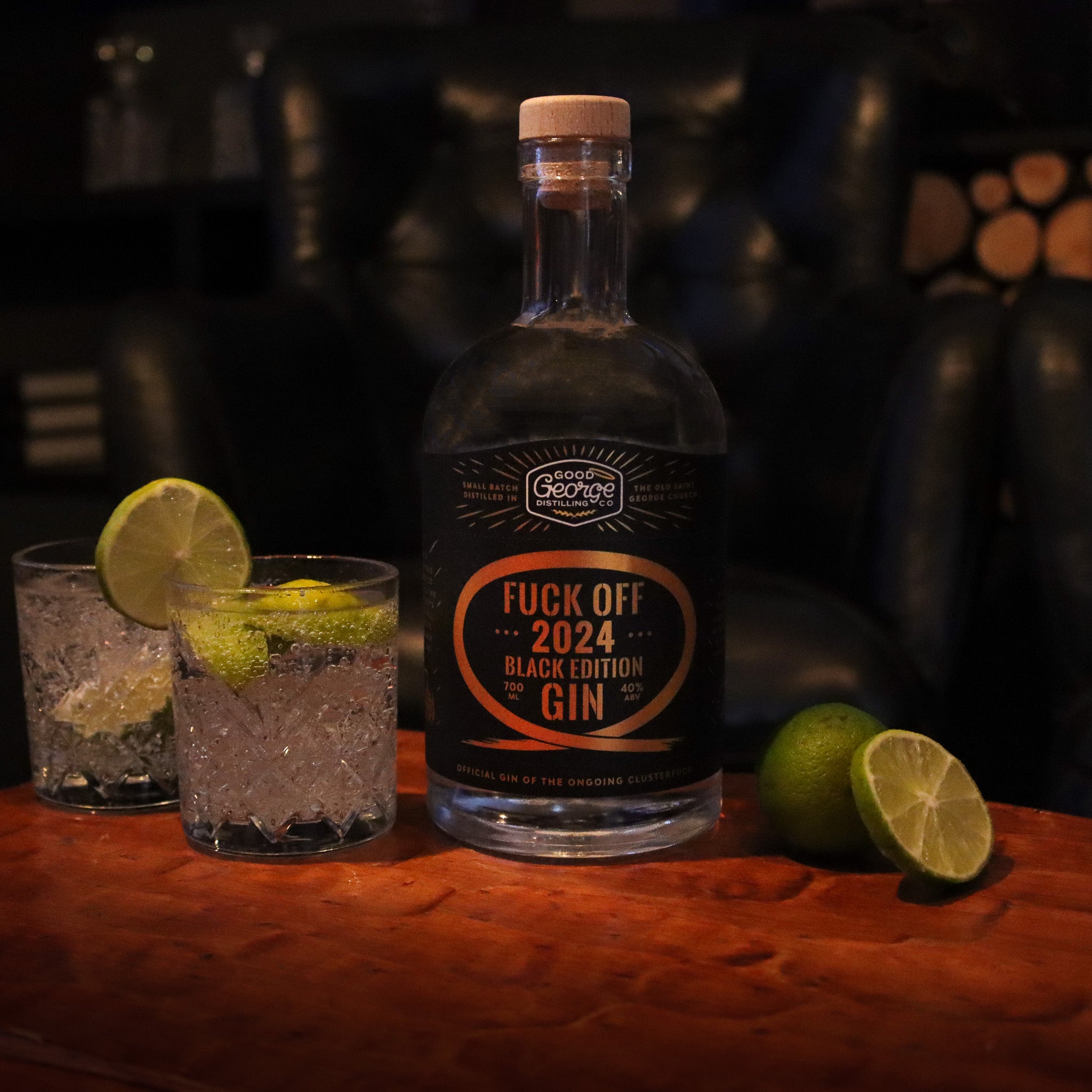 A bottle of Fuck Off 2024 Black Edition Gin with a label that has gold and orange text on a dark background. A gin and tonic next to it and fresh cut limes