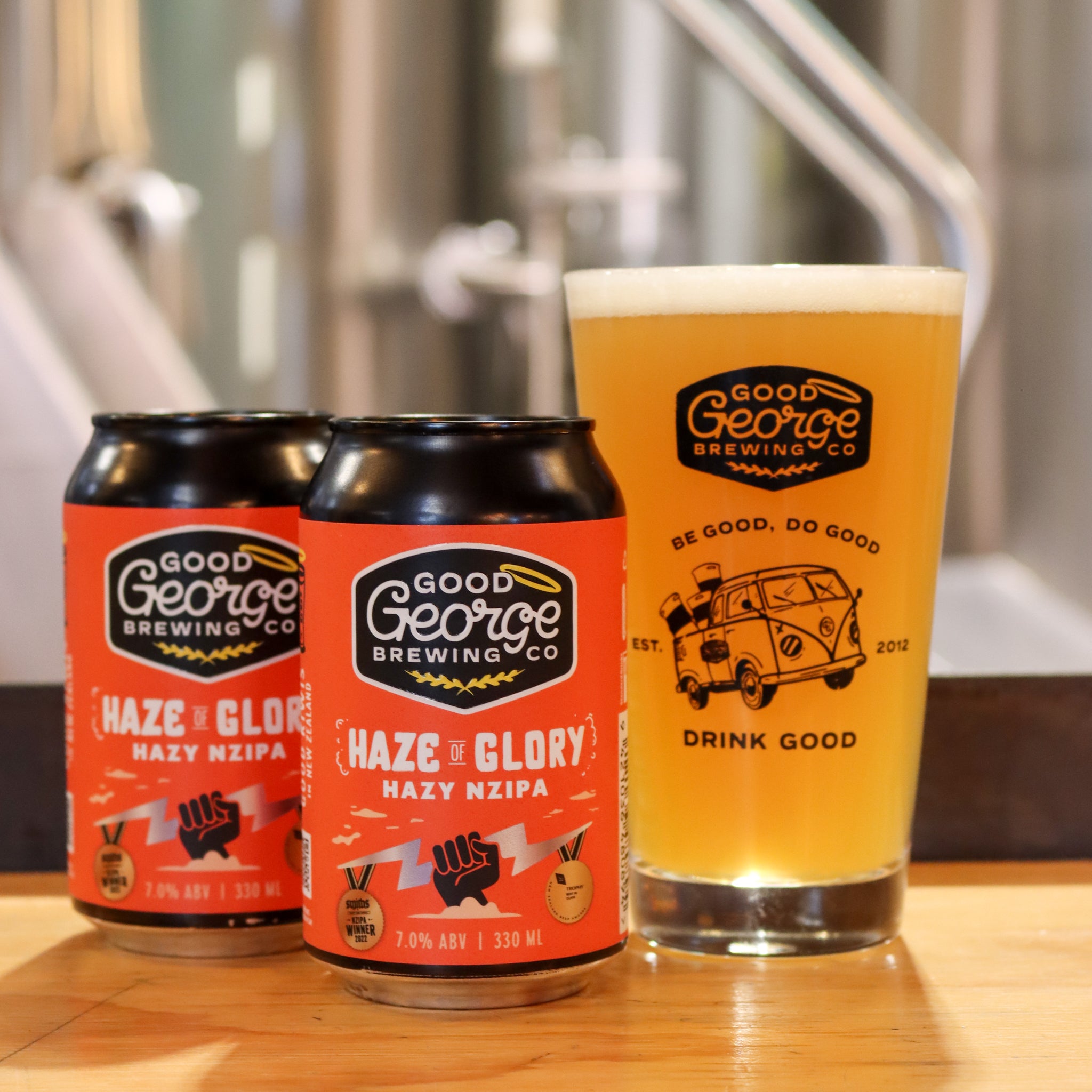 Two cans and a glass of Good George Brewing Co. beer on a wooden surface.