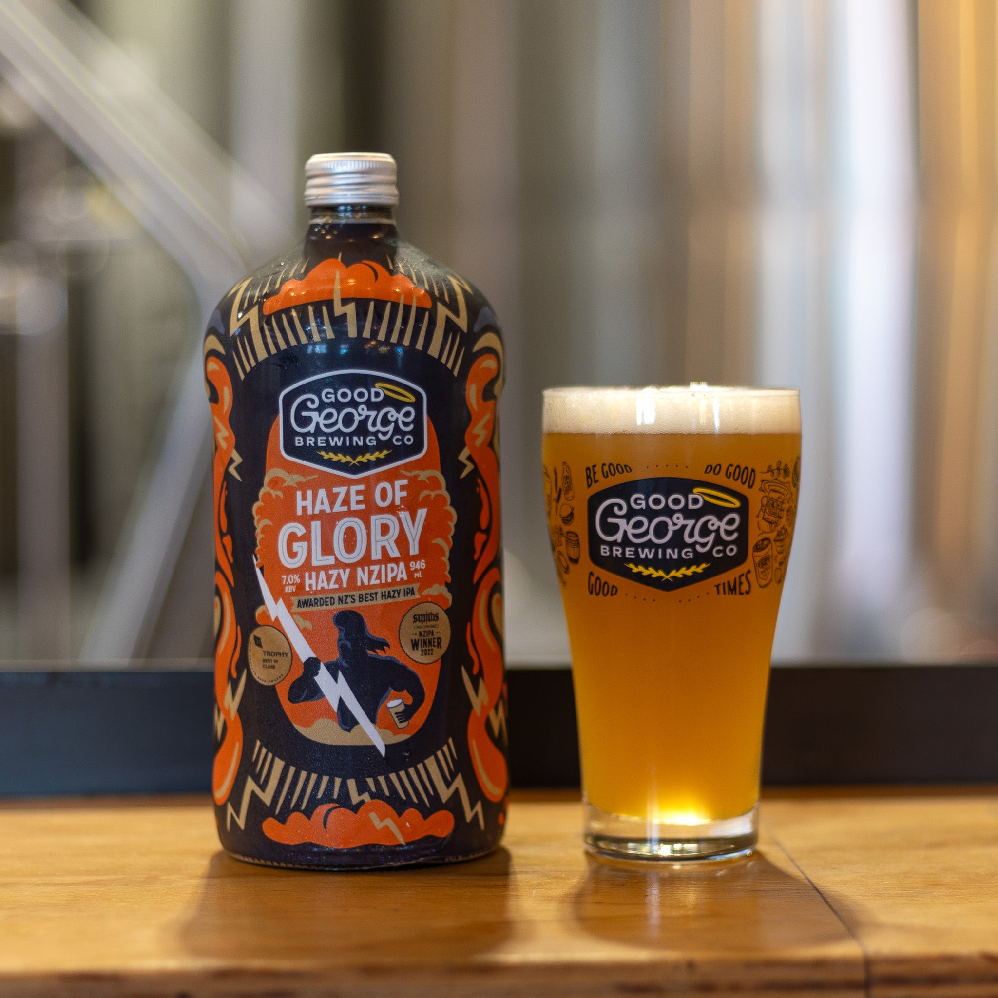 A bottle of Haze of Glory Hazy NZIPA beer with a full beer Good George Beer Glass next to with the brewery blurred in the background