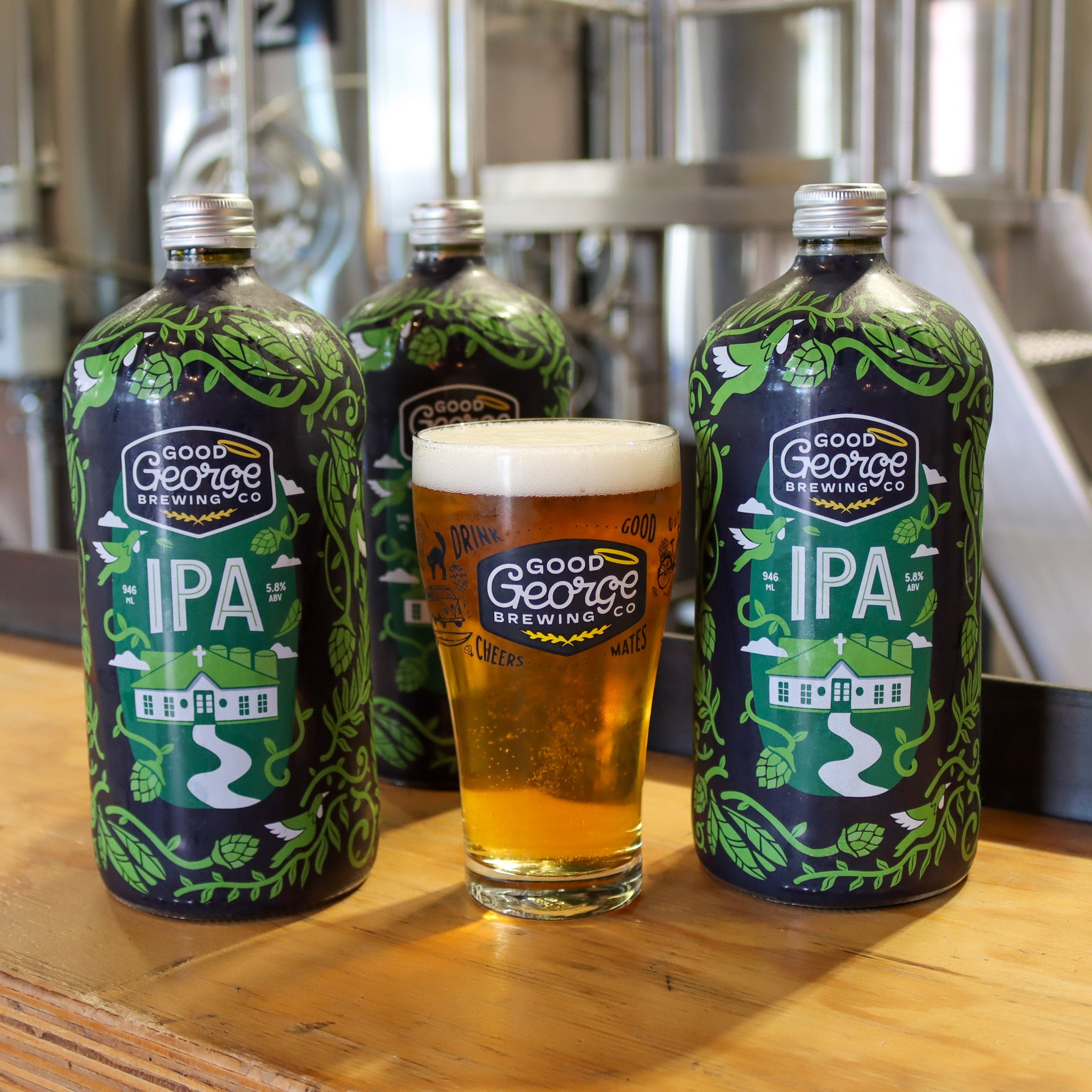 3 bottles of IPA beer with a full Good George Beer Glass next to with the brewery blurred in the background