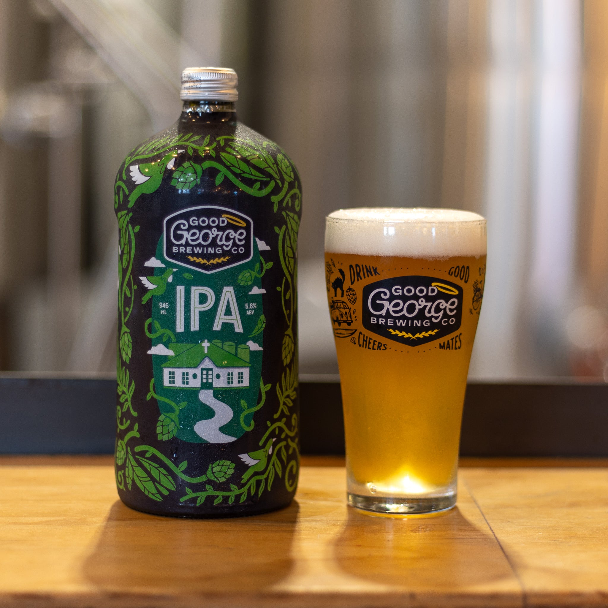 A bottle of IPA beer with a full Good George Beer Glass next to it with the brewery blurred in the background