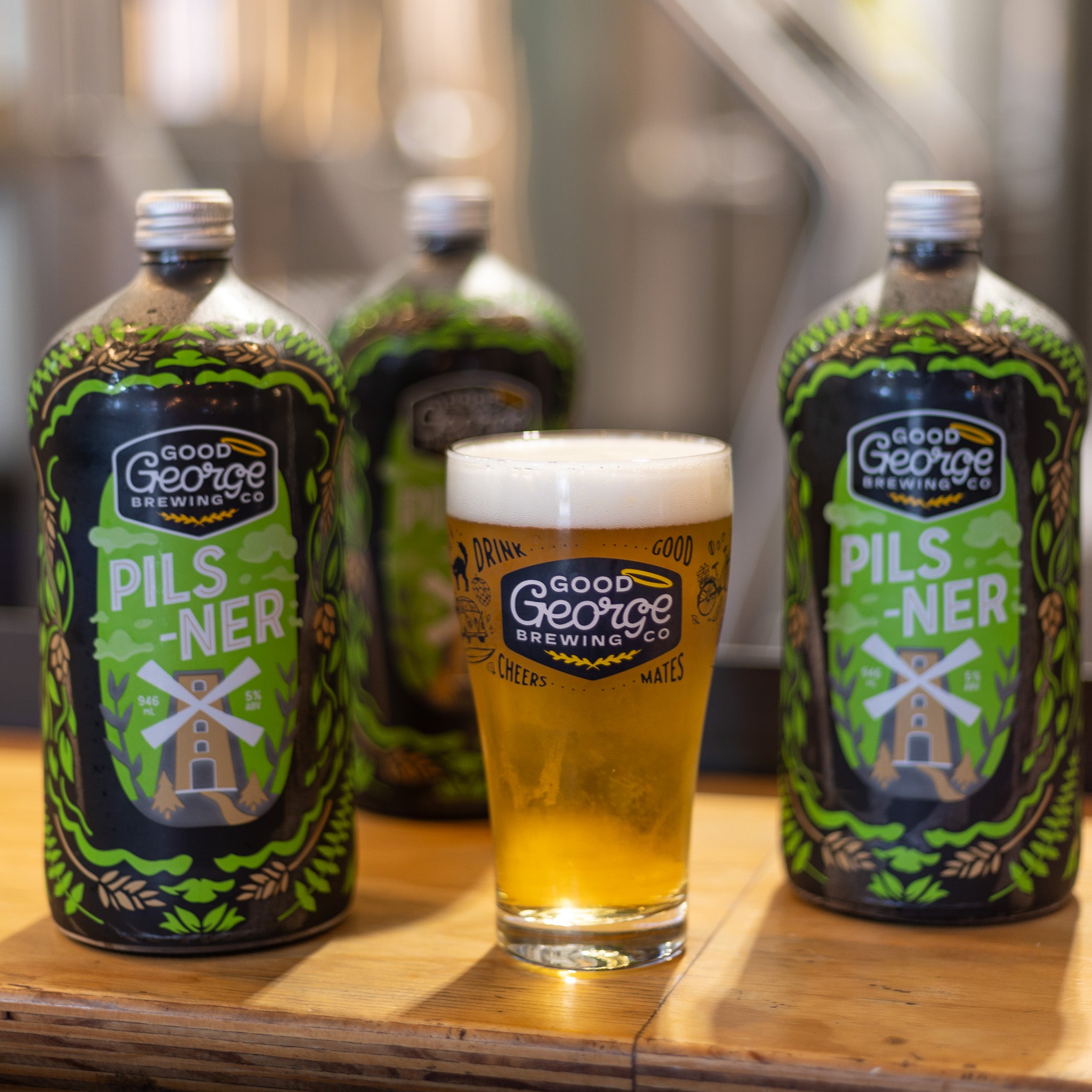3 bottles of Pilsner beer with a full Good George Beer Glass next to with the brewery blurred in the background