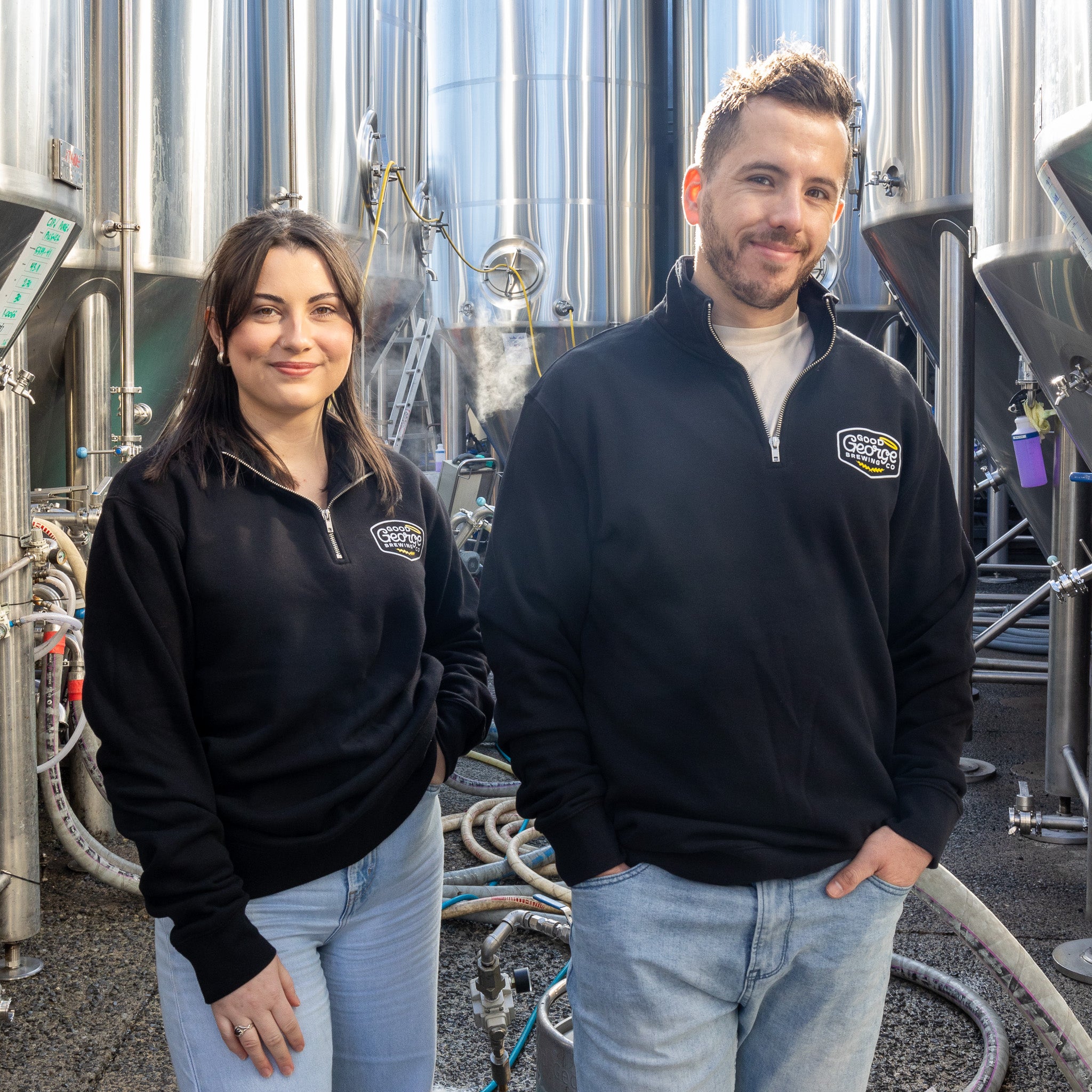 Two individuals standing in front of industrial equipment, wearing black quarter zip sweaters with the 'Good George' brand logo.