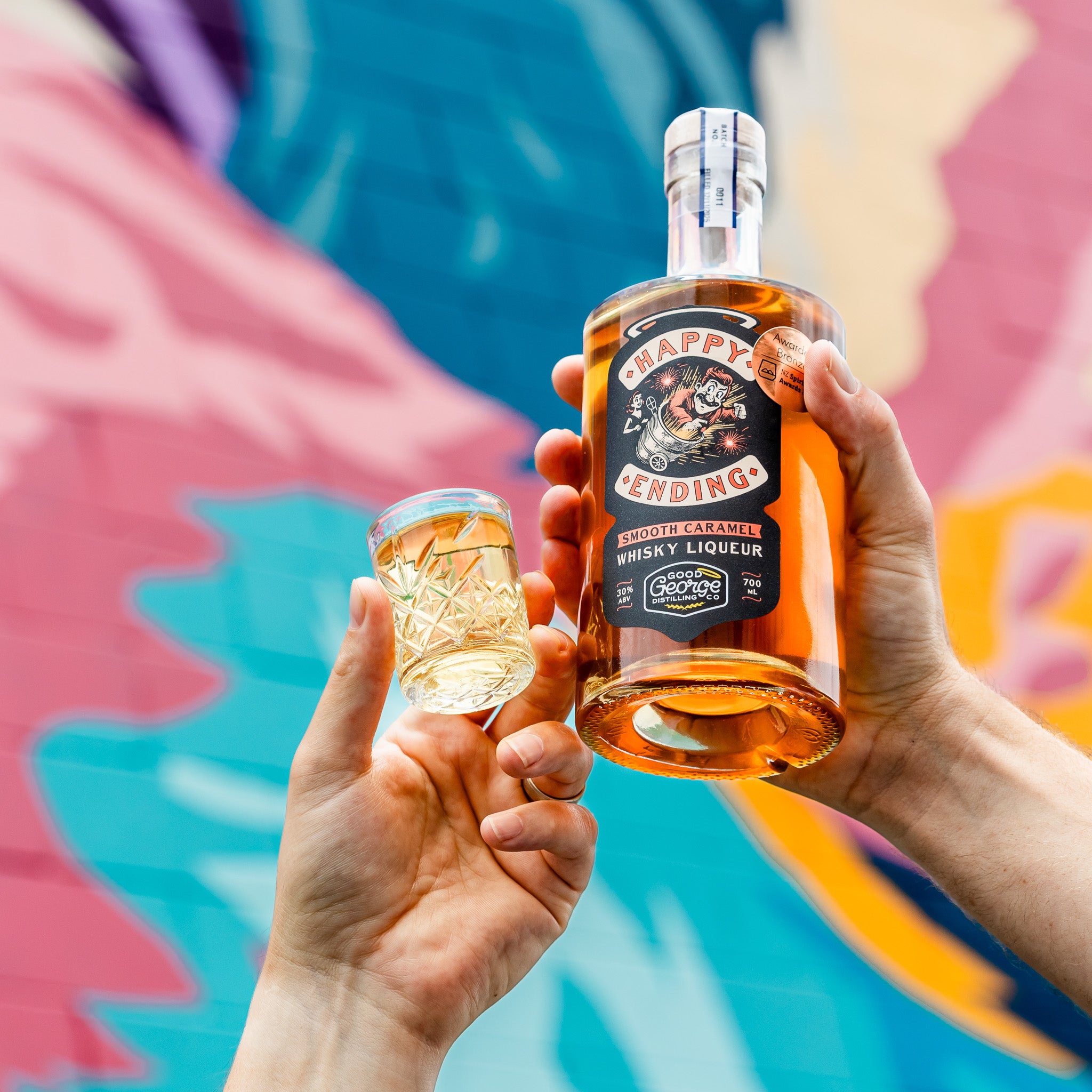 Two hands holding a bottle and a glass of 'Happy Ending' whiskey liqueur against a colorful abstract background.