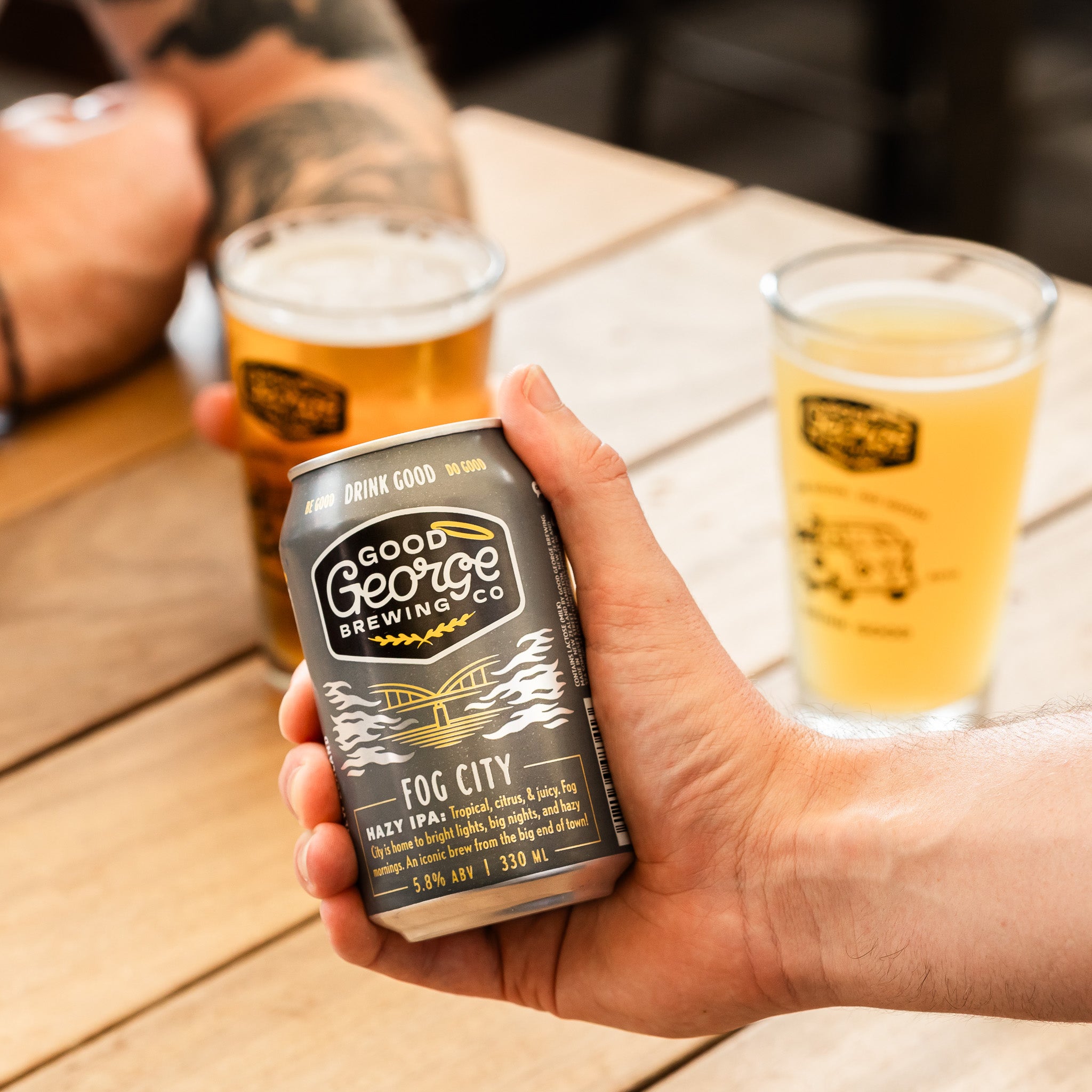 Hand holding a can of Good George Brewing Co. Fog City beer with two glasses of beer in the background.