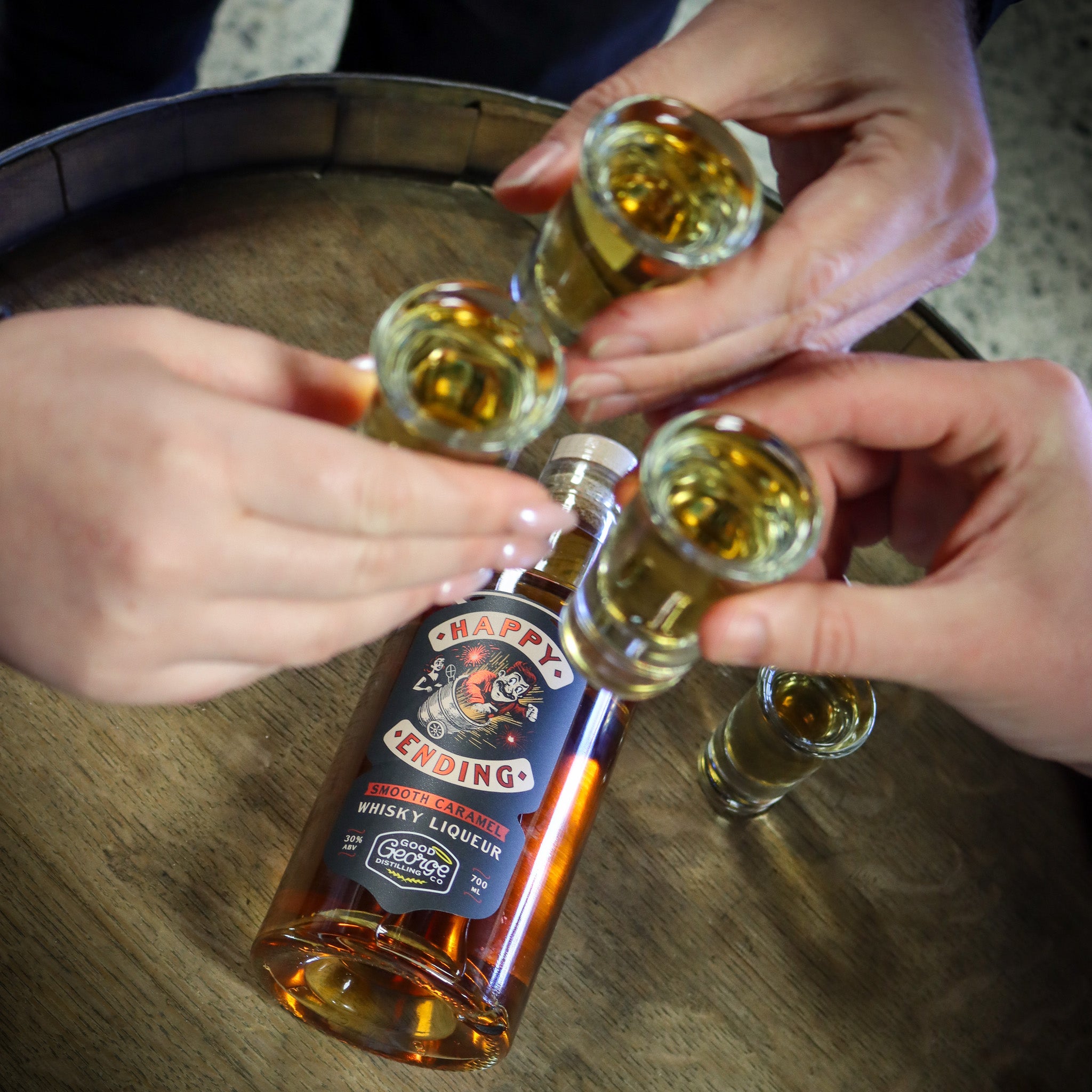 Four hands holding shot glasses with a bottle of Happy Ending whisky liqueur on a barrel.