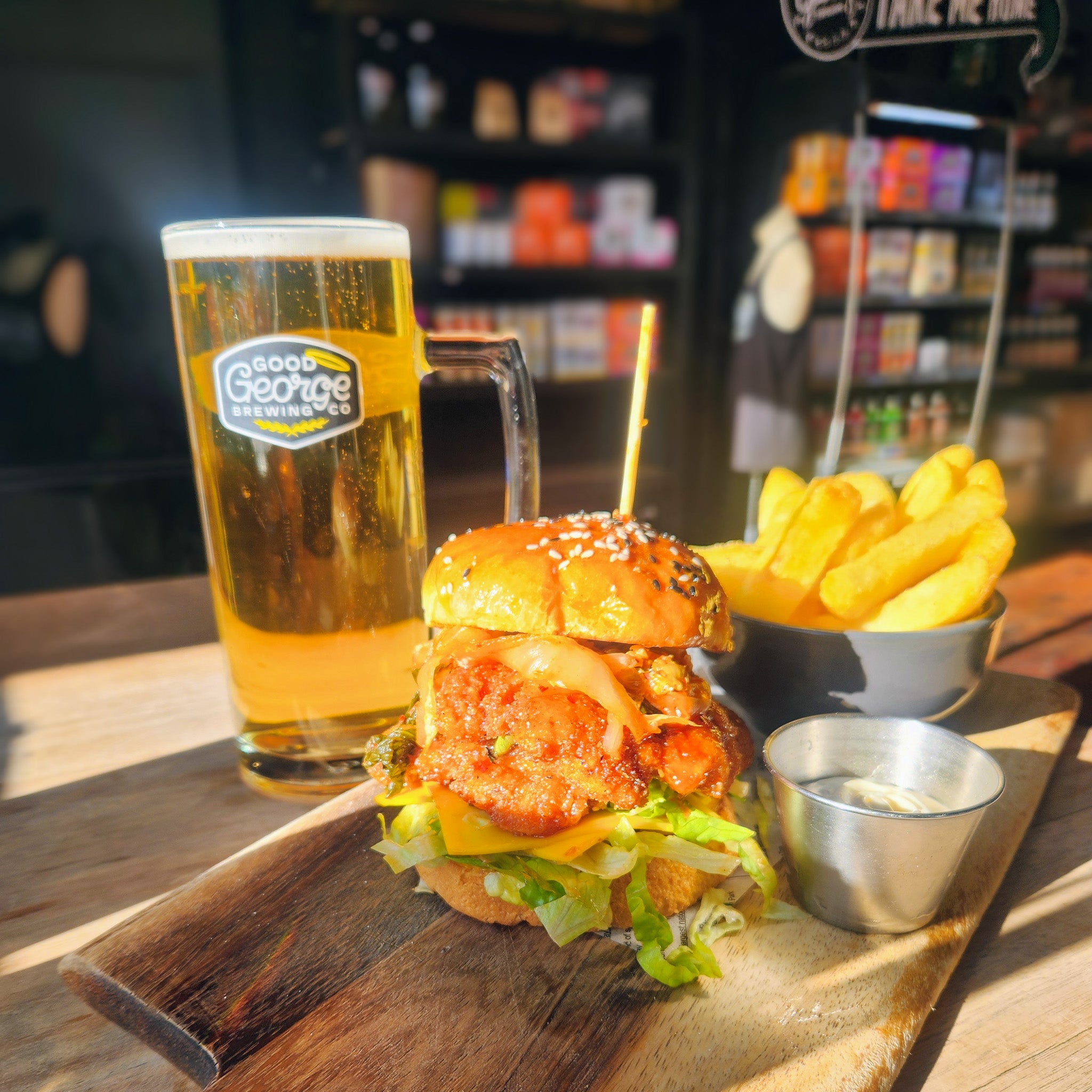 A large burger with a thick breaded patty, lettuce, and onion, served on a wooden board with a glass of beer and a side of fries in a pub setting.