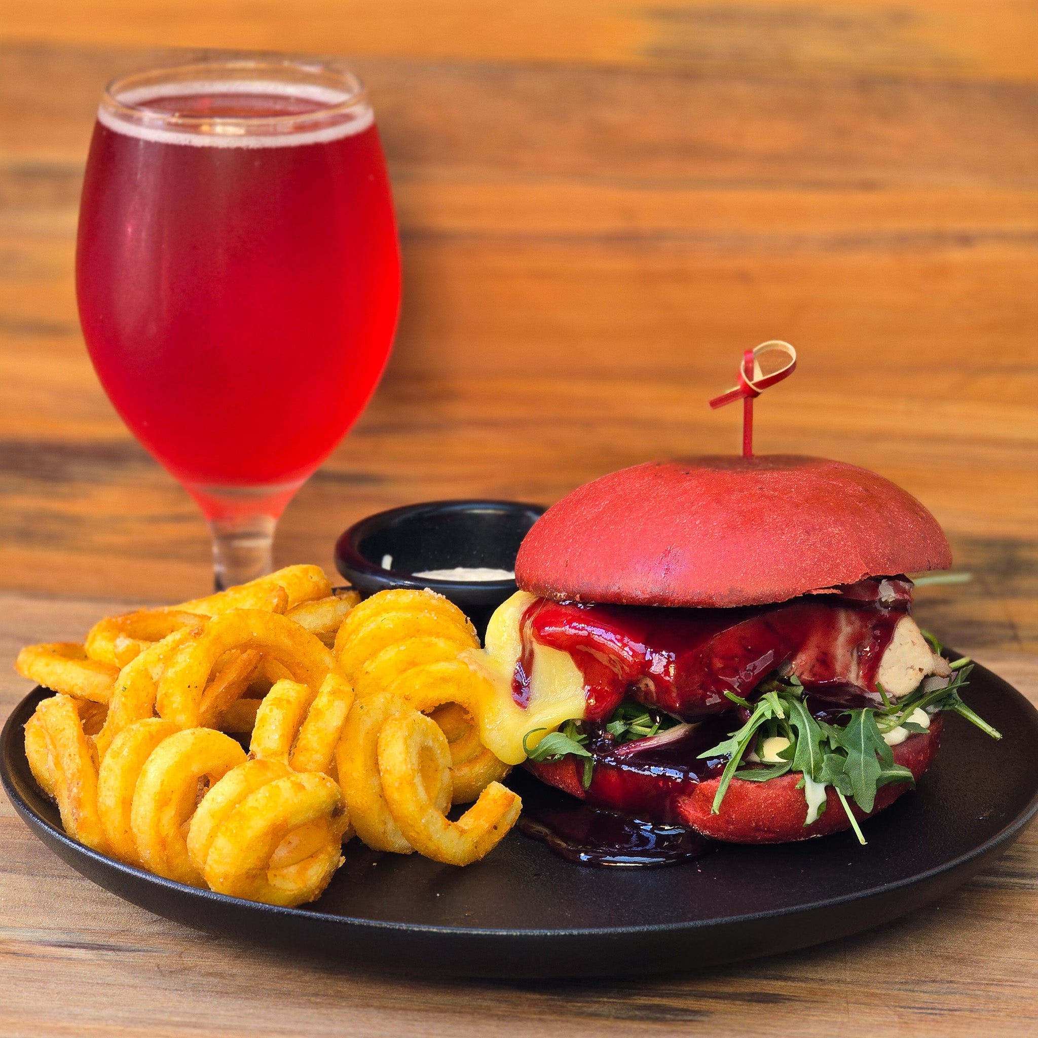 A plated burger with a bright red bun, dark red sauce, greens, and a curly fries side, served on a wooden table with a glass of red beverage.