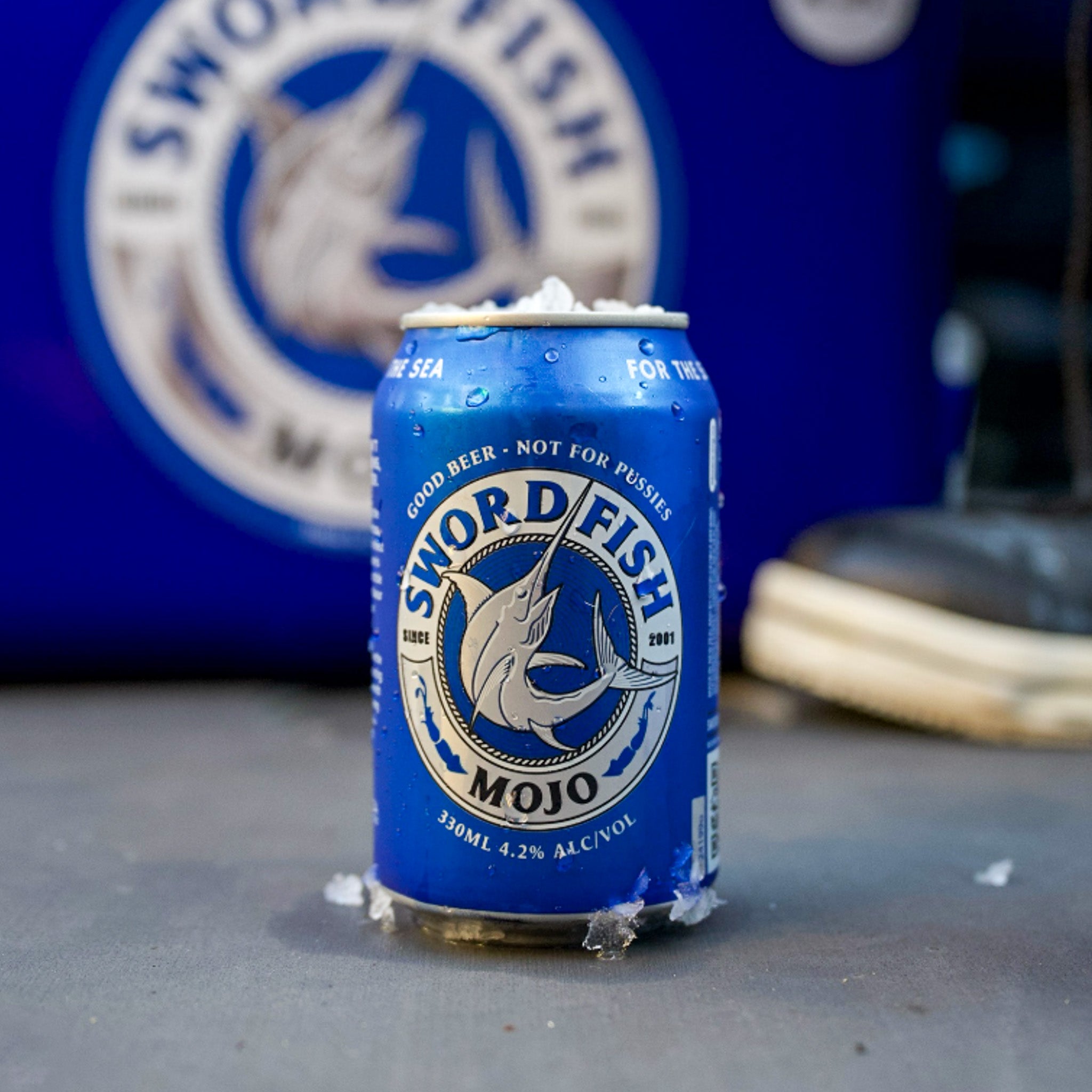 A blue can of Sword Fish Mojo beer sits on a gray surface with condensation droplets, set against a blurred blue background featuring the brand's logo and a shoe.