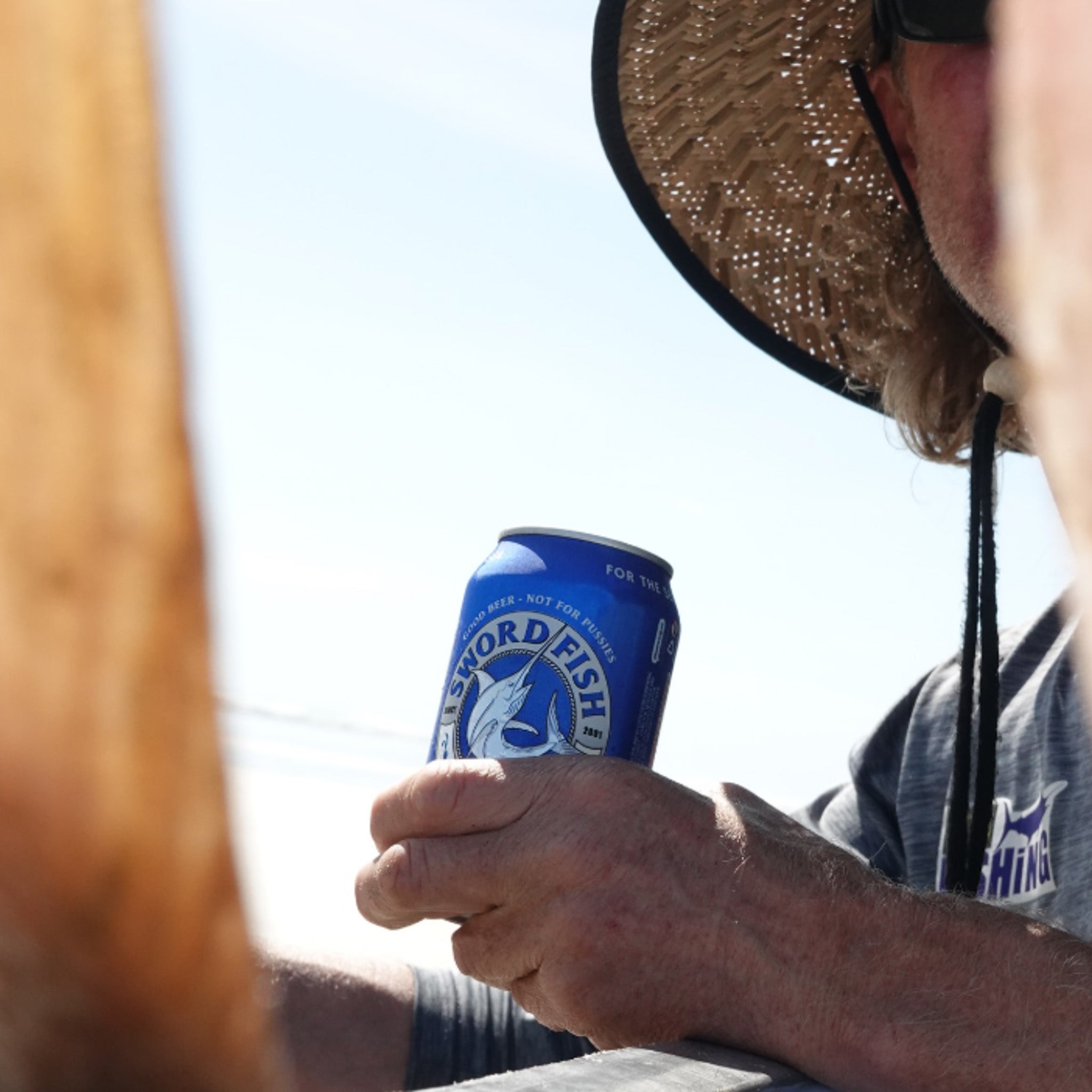 A person wearing a straw hat and sunglasses holds a blue can of Sword Fish Mojo beer outdoors against a bright sky.