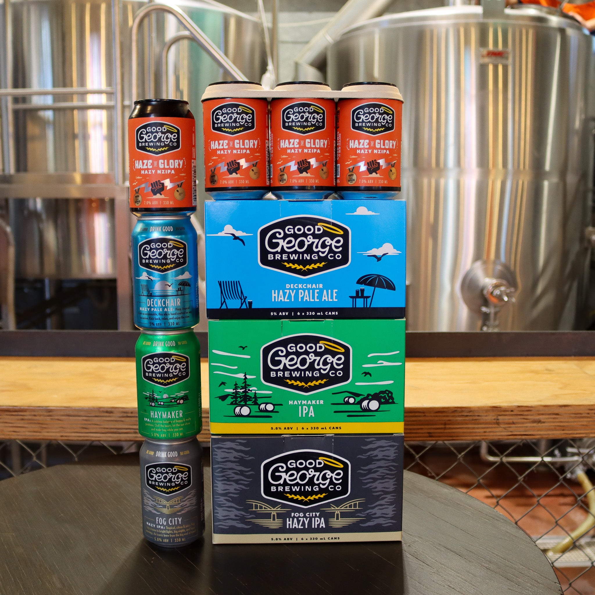 Stack of Good George Brewing Co. beer cans and boxes on a table with brewing equipment in the background.