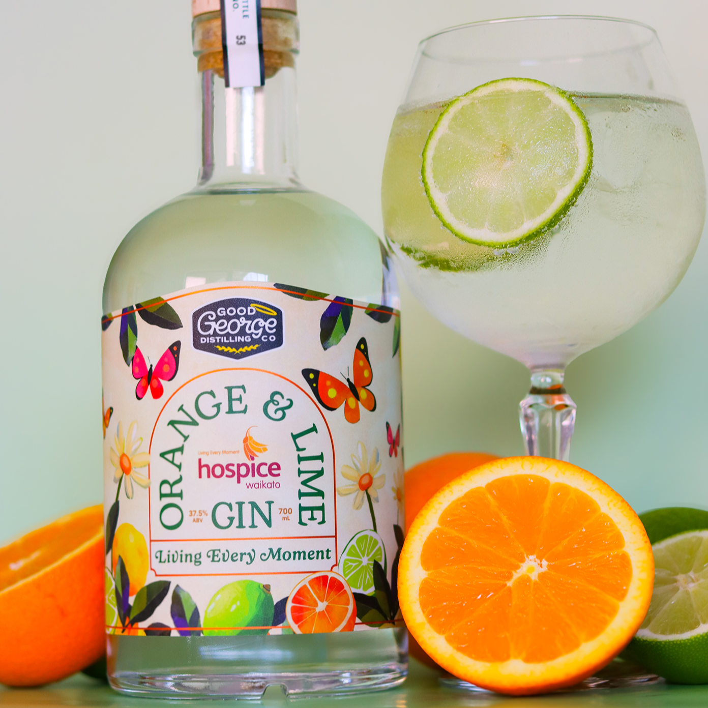 A bottle of Hospice Waikato 'Living Every Moment' Orange & Lime Gin with a glass of gin and tonic, orange slices, and a lime slice in the foreground.