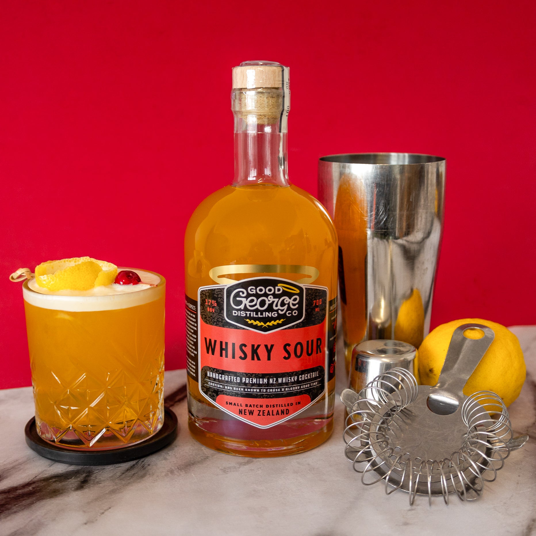 A bottle of Good George Whisky Sour with a yellow label, featuring text about the product being a handcrafted cocktail made with 5 year old NZ Single Malt Whisky and a whisky sour cocktail next to it and a cocktail shaker