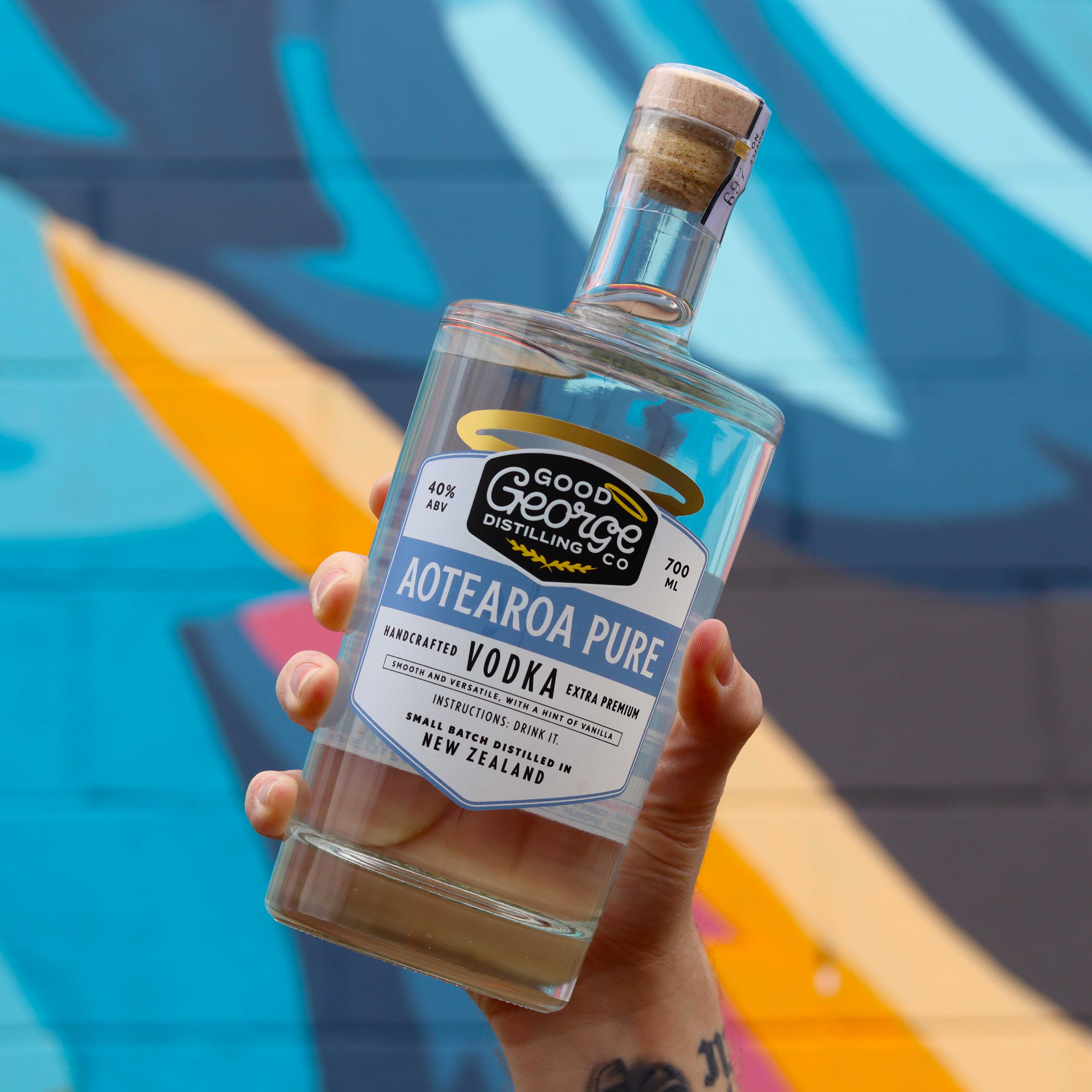 A bottle of Aotearoa Pure Vodka by Good George Distilling Co. with a white label and gold accents on a blue background holded up in front of a mural.