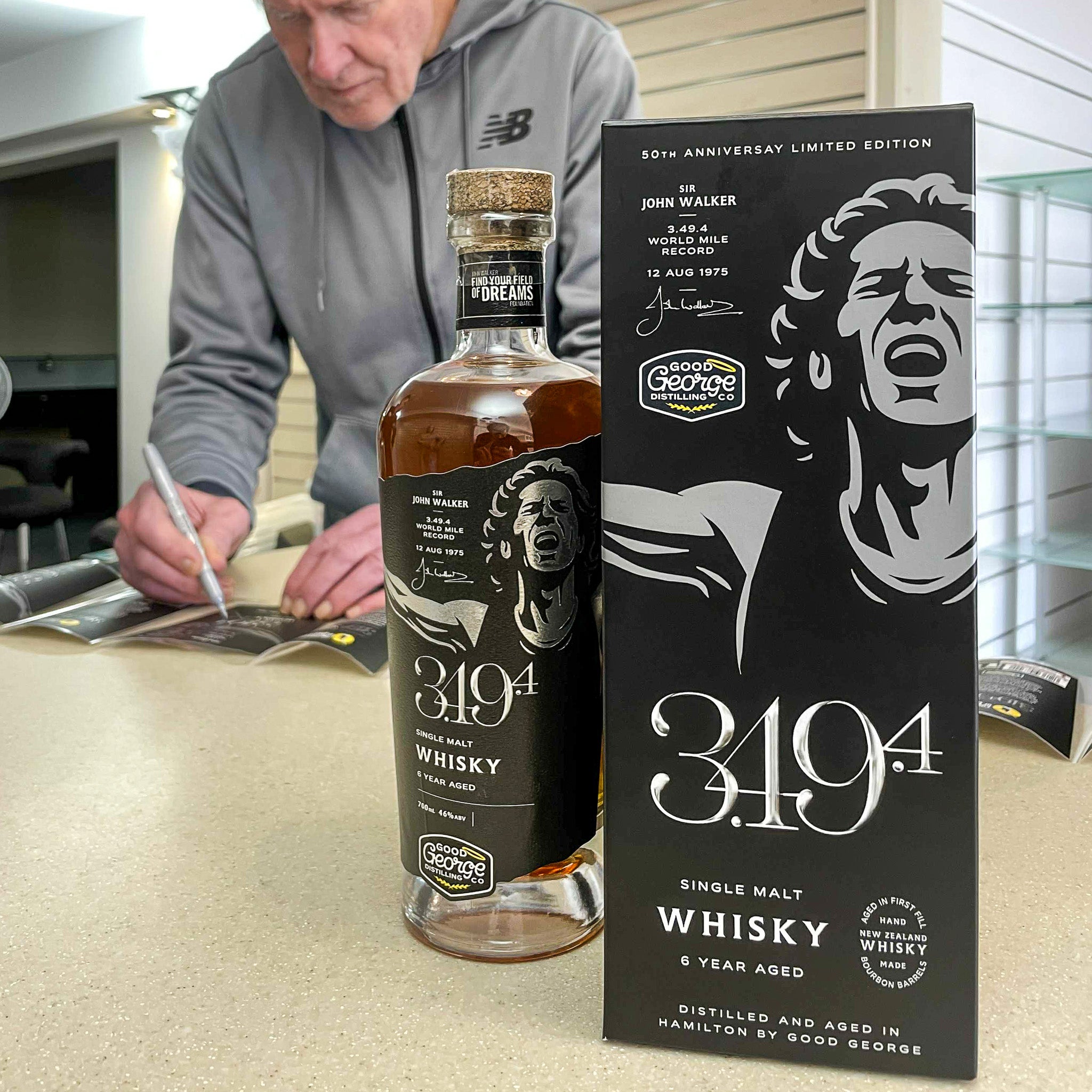 John Walker's whisky bottle and packaging on a table with John Walker signing some labels in the background.