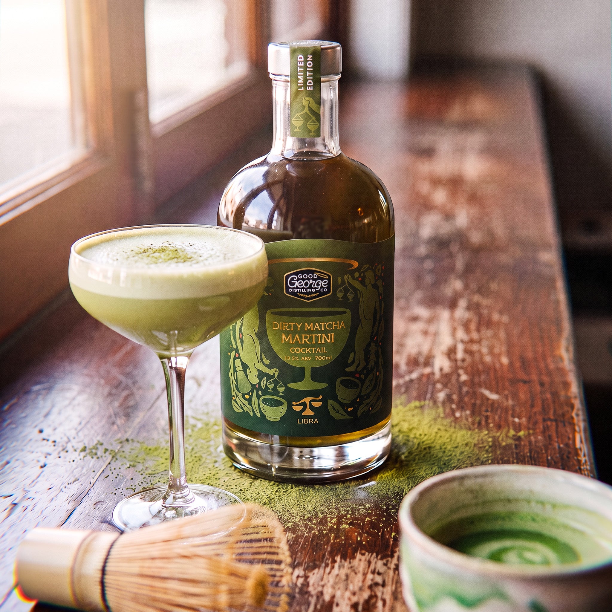 A bottle of Libra Matcha Martini Cocktail next to a prepared matcha latte in a coupe glass, with a whisk and ceramic cup on a rustic wooden table near a window.