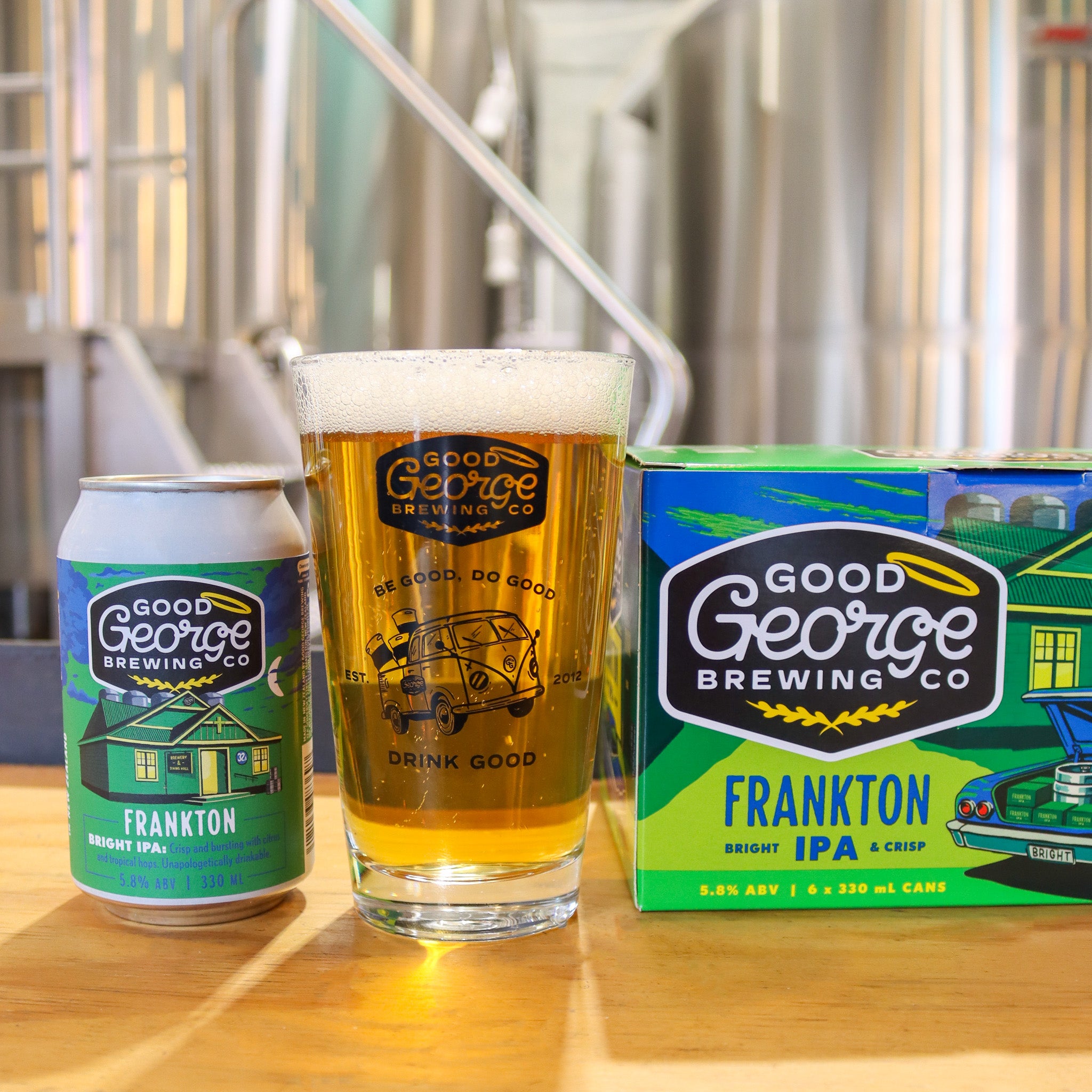 Good George Brewing Co. Frankton IPA can, glass, and box on a wooden surface with brewing equipment in the background.