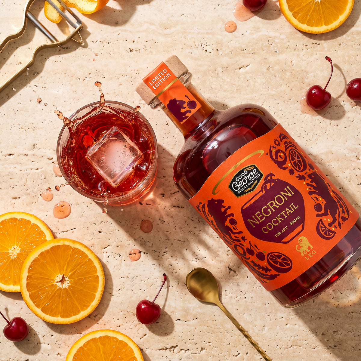 A bottle of Good George Negroni Cocktail liqueur and a glass of the red cocktail with an ice cube, styled on a light stone surface with orange slices, cherries, a spoon, and a peeler.