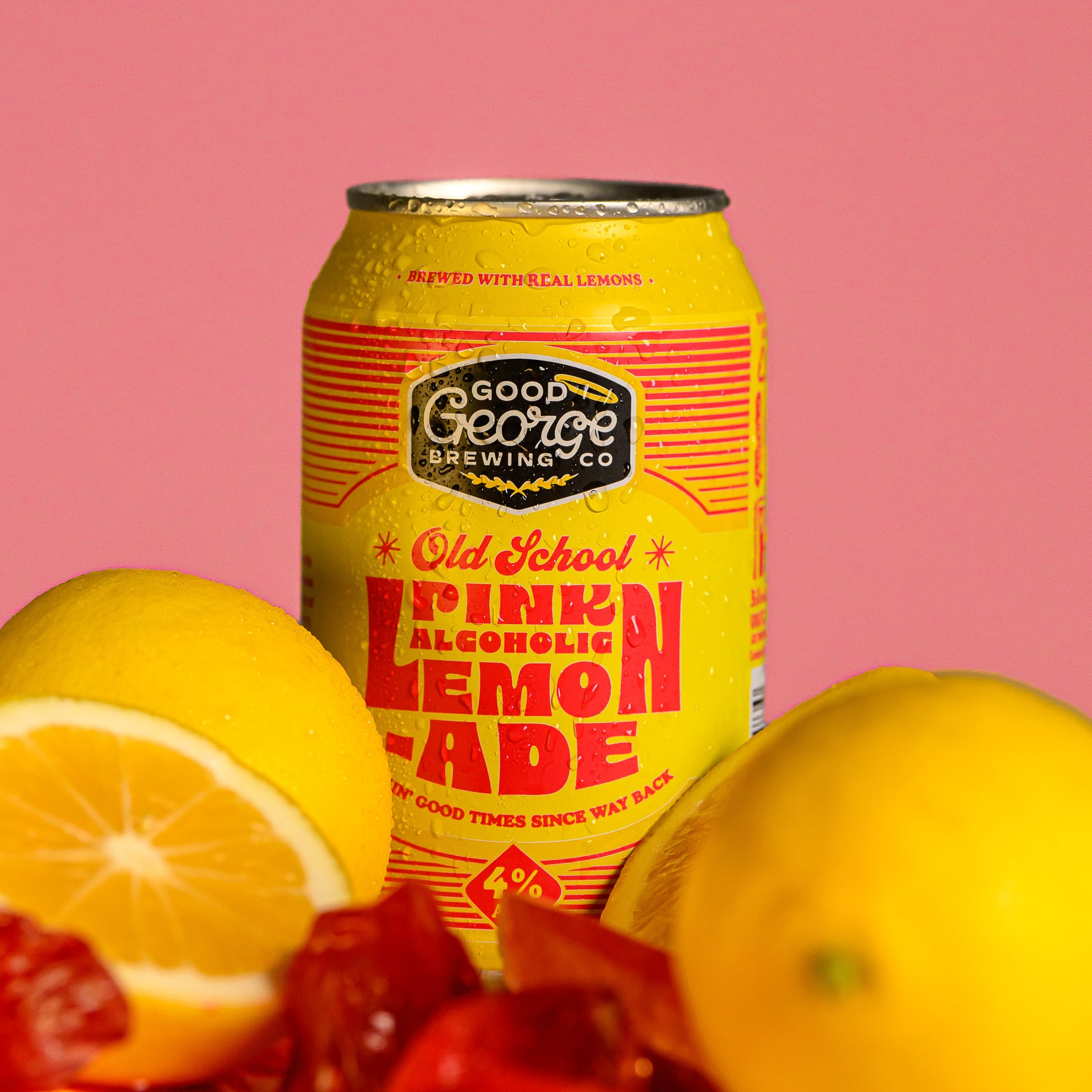 A yellow can of Good George Brewing Co. Old School Pink Lemonade is centered against a solid pink background, surrounded by fresh lemons and strawberries in the foreground.