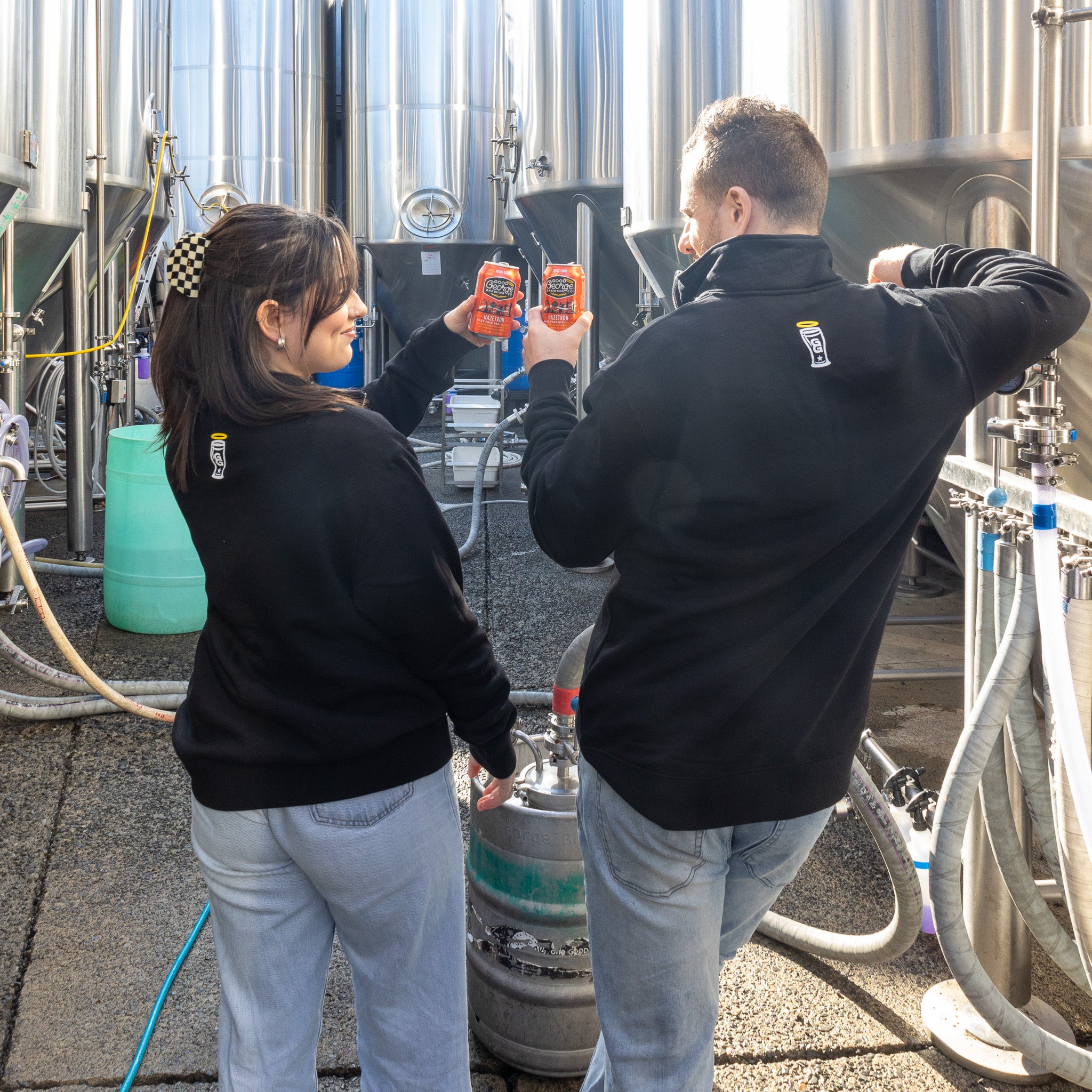 Two individuals standing in front of industrial equipment, wearing black quarter zip sweaters with the 'Good George' brand logo.