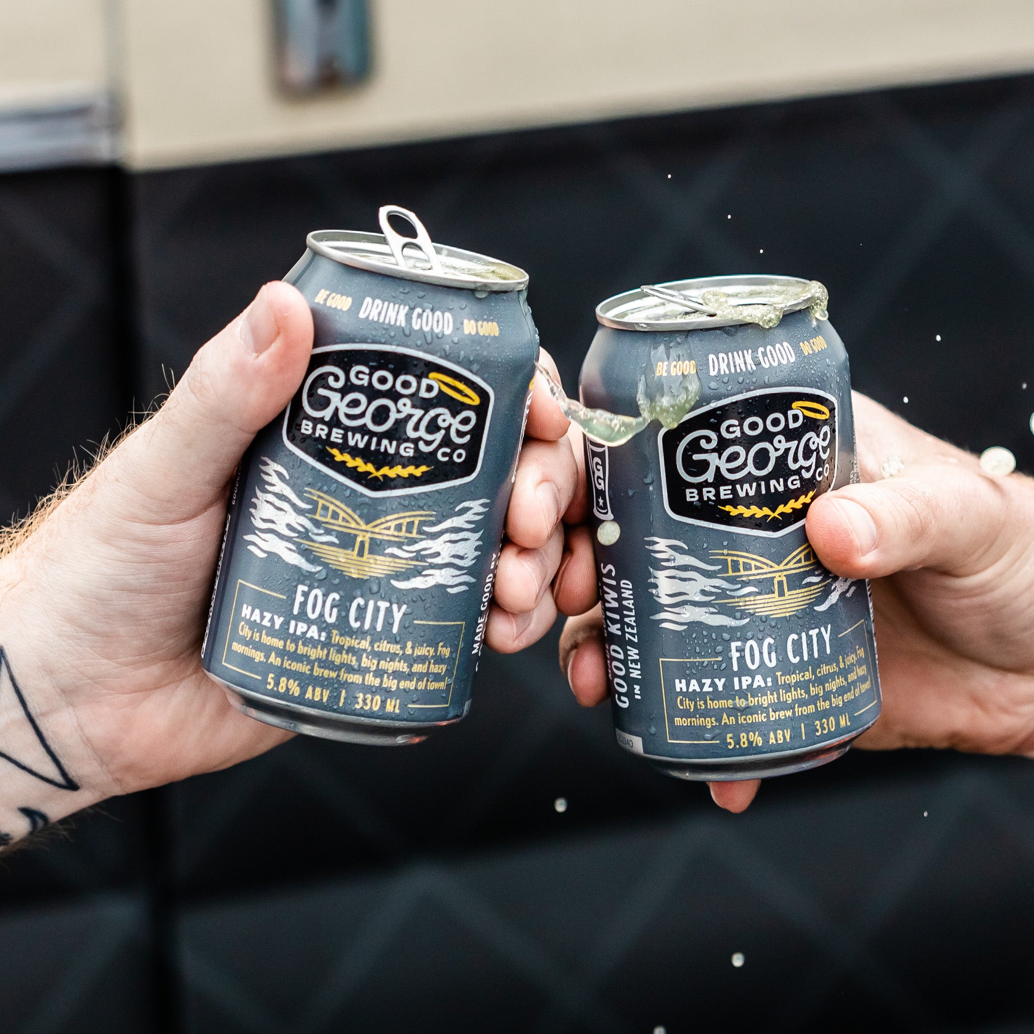 Two cans of Good George Brewing Co. Fog City beer being held up against a dark background.