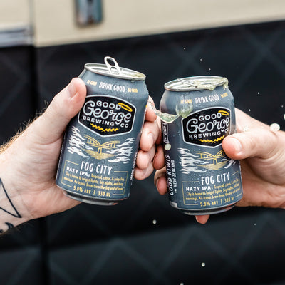 Two cans of Good George Brewing Co. Fog City beer being held up against a dark background.