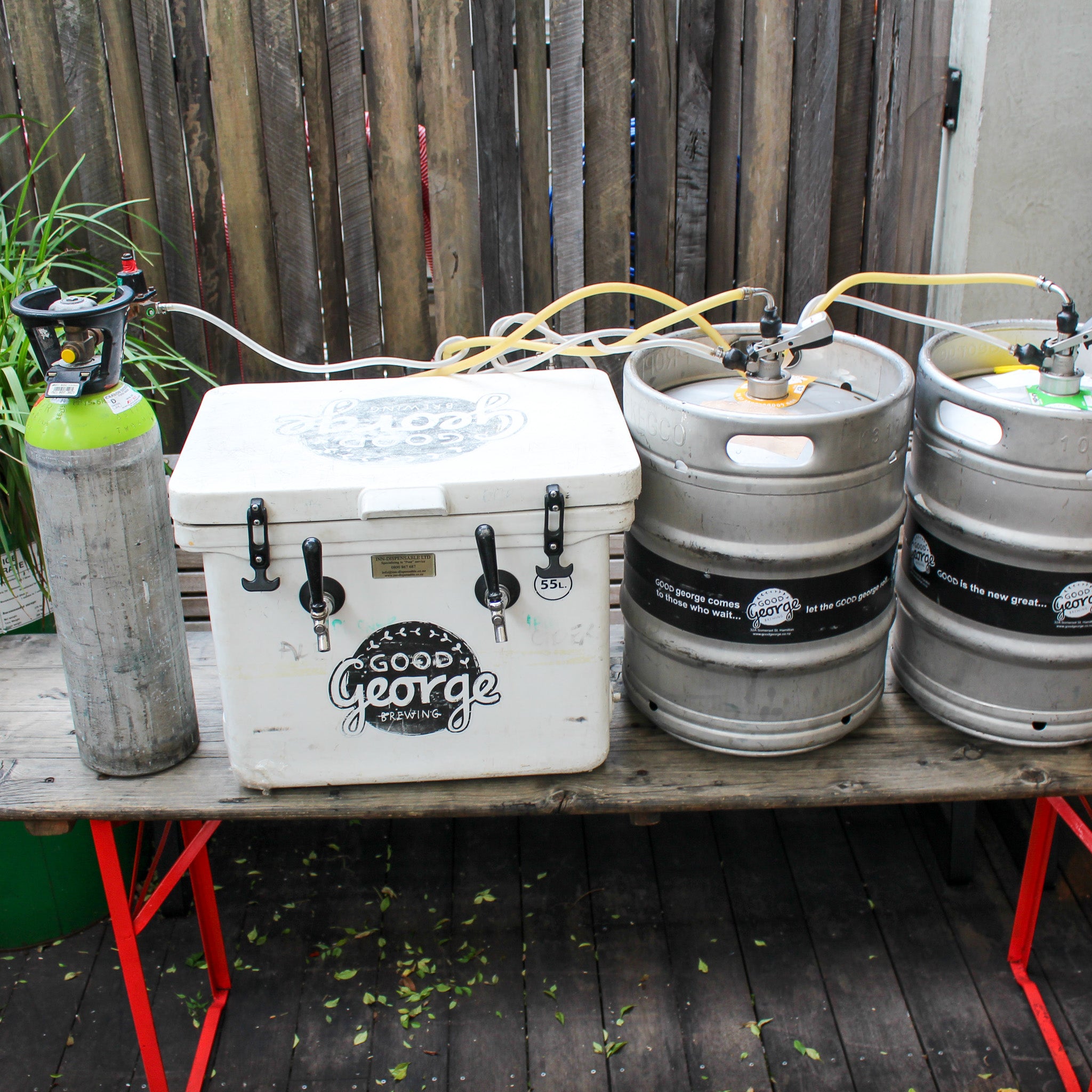 Cooler with beer taps next to kegs and a gas tank on a wooden table outdoors.
