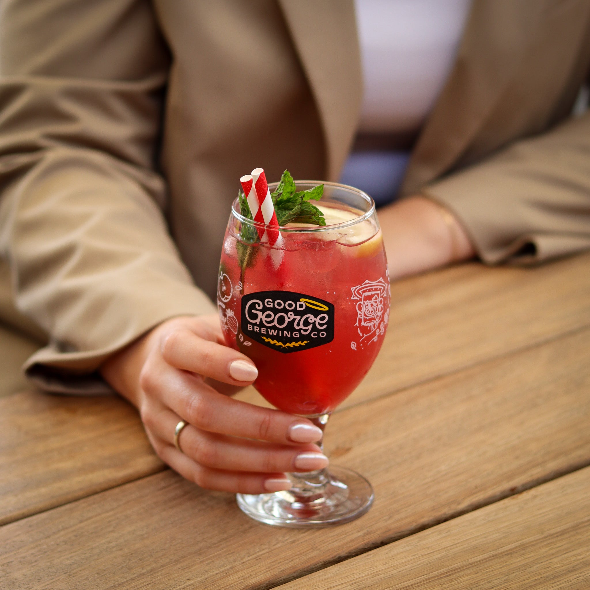 Person holding a red cocktail in a glass with 'Good George Brewing Co' label on a wooden table.