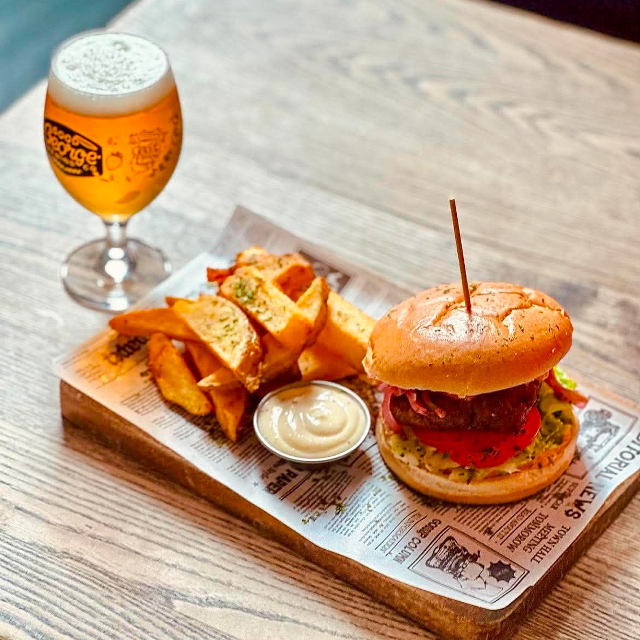 A burger with a wooden toothpick, served on a newspaper-lined wooden board with potato chips and a small dipping sauce, next to a glass of beer on a wooden table.
