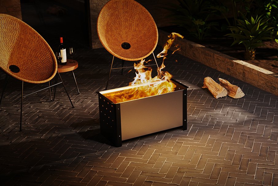 A rectangular metal fire pit with a warm metallic finish and black sides, shown lit with flames on a dark brick patio. Two woven accent chairs and a small side table are visible in the background.