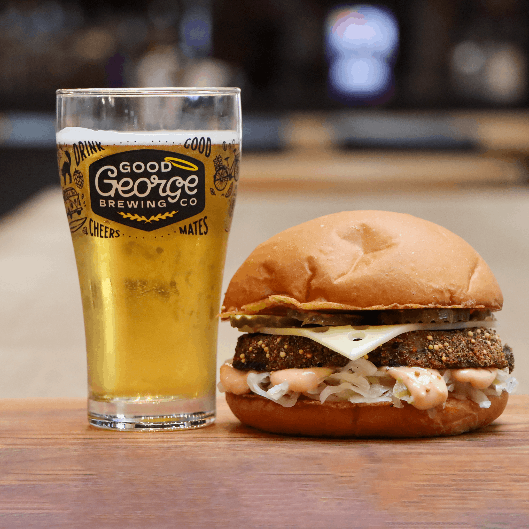 A beer glass and a burger on a wooden table in a bar setting. The beer glass is branded with 'Good George Brewing Co'.