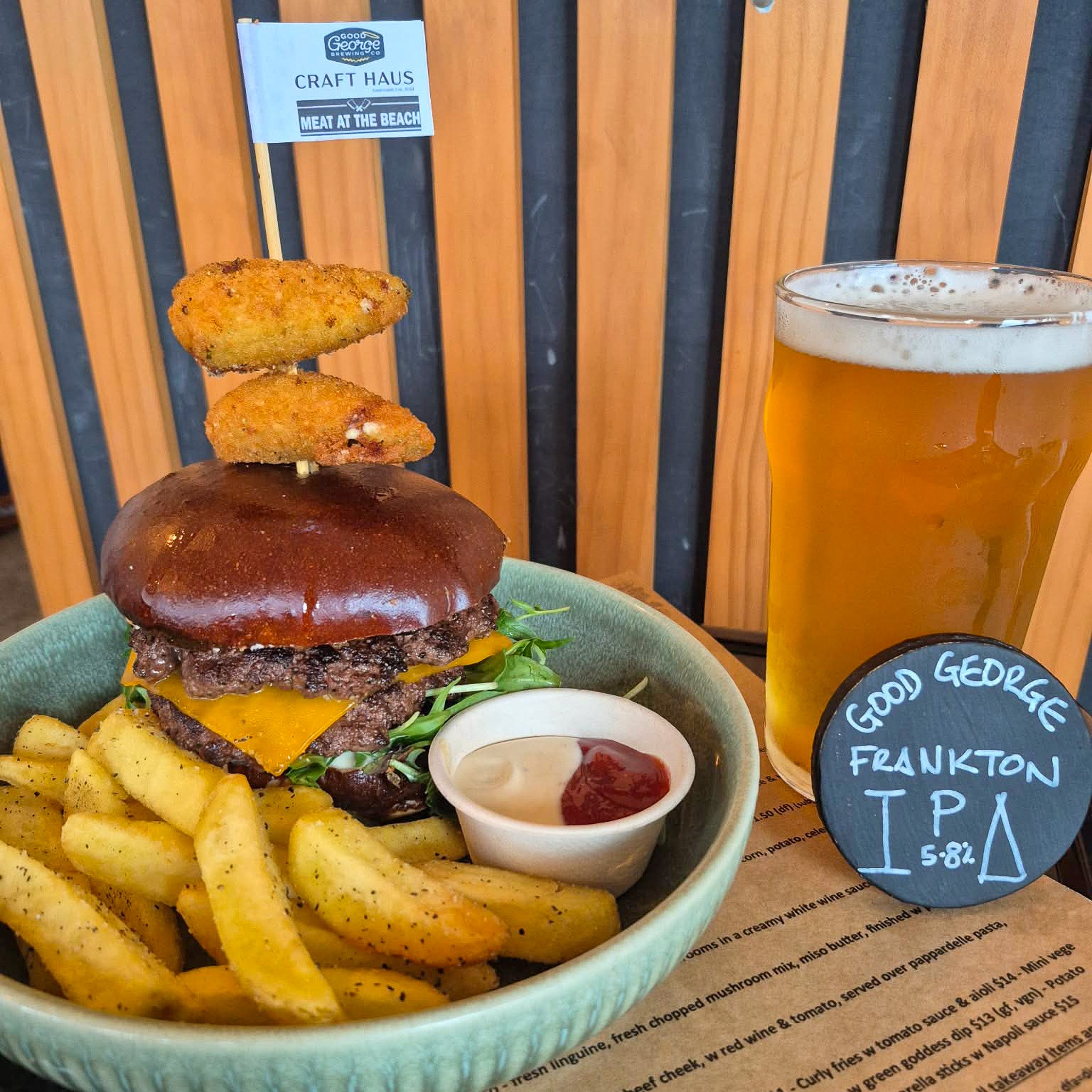 A burger served in a light blue bowl with golden fries, a small cup of tartar sauce, and a glass of beer on a restaurant table. The burger has a dark brown sauce on the bun, cheese, beef patties, and two fried onion rings stacked on top.