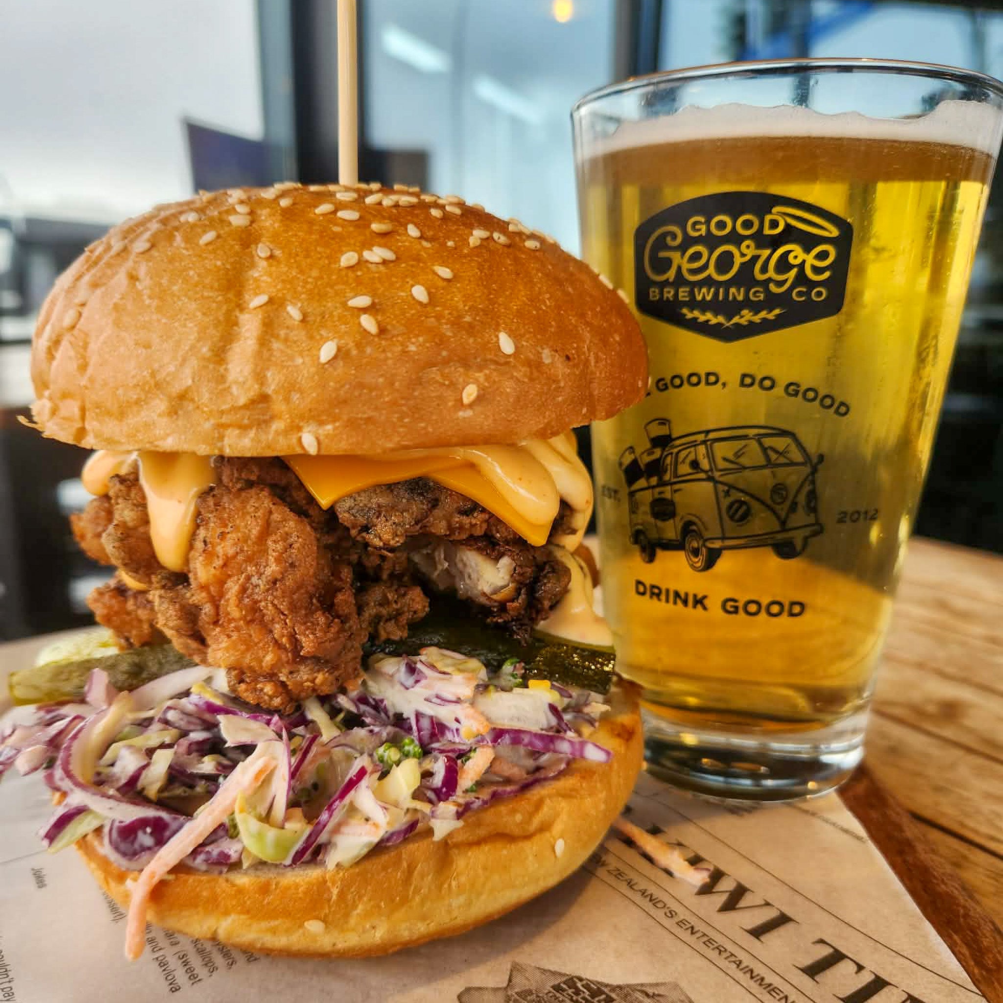A close-up of a large gourmet burger with sesame buns, fried chicken, melted cheese, coleslaw, and pickles, served next to a clear glass of golden beer printed with the Good George Brewing Co logo on a wooden table.