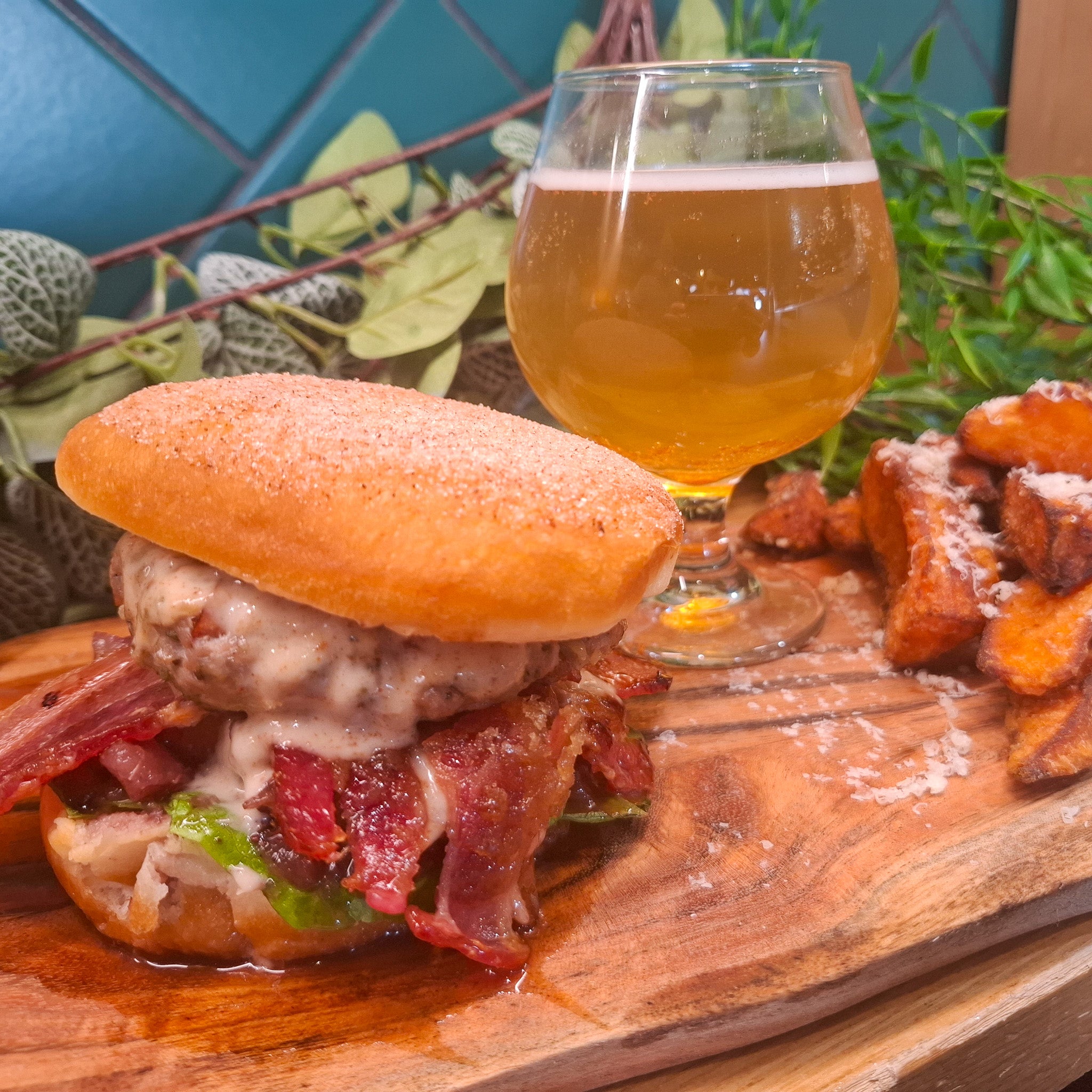 A close-up food photo of a loaded breakfast sandwich on a wooden board, served with a glass of beer and potato wedges. The sandwich contains a sausage patty, melted cheese, bacon, and green peppers in a bun dusted with sugar.