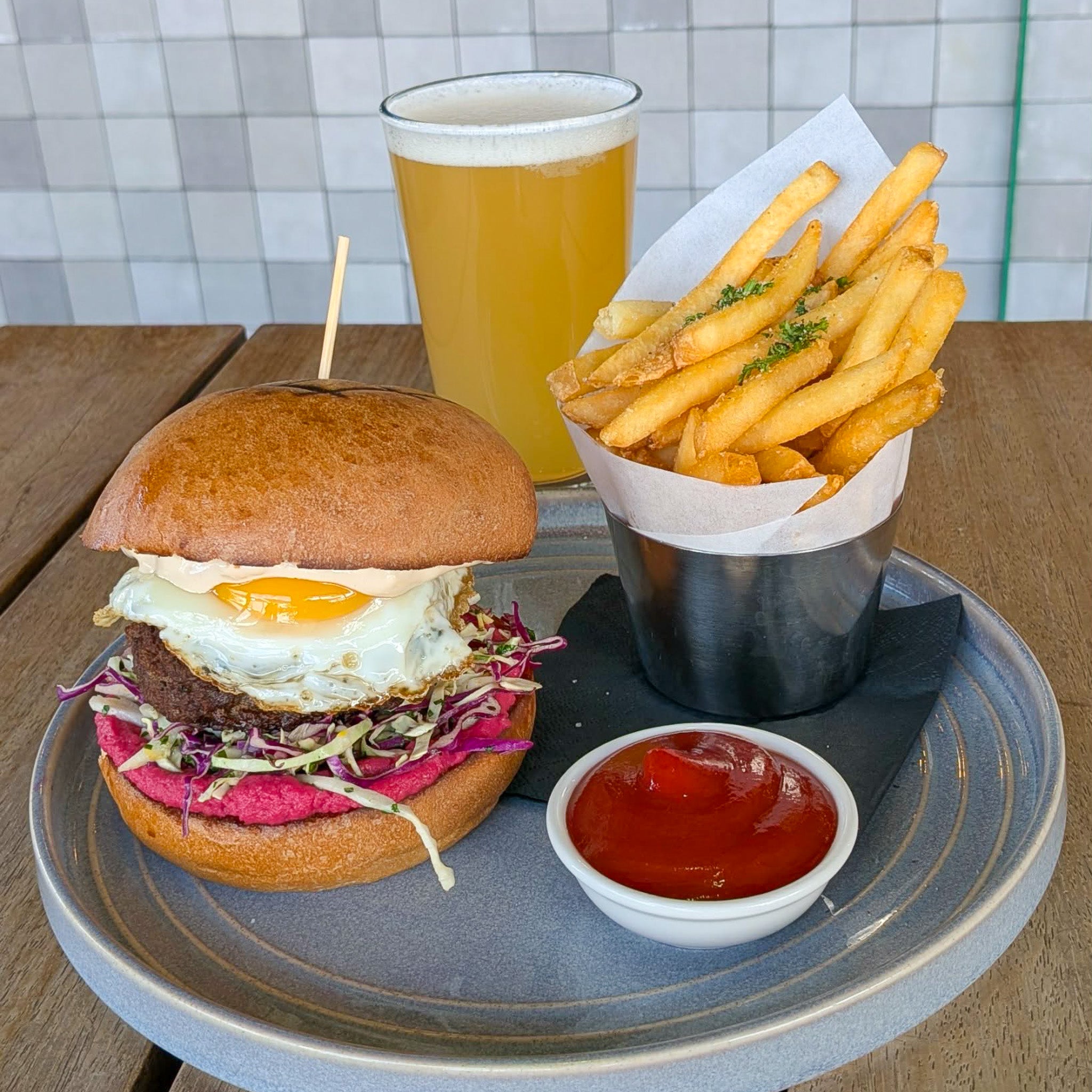 A burger with a fried egg, patty, and shredded cabbage on a gray plate, served with a metal bucket of french fries and a small dish of ketchup. A glass of beer stands behind the plate on a wooden table with a tiled wall in the background.