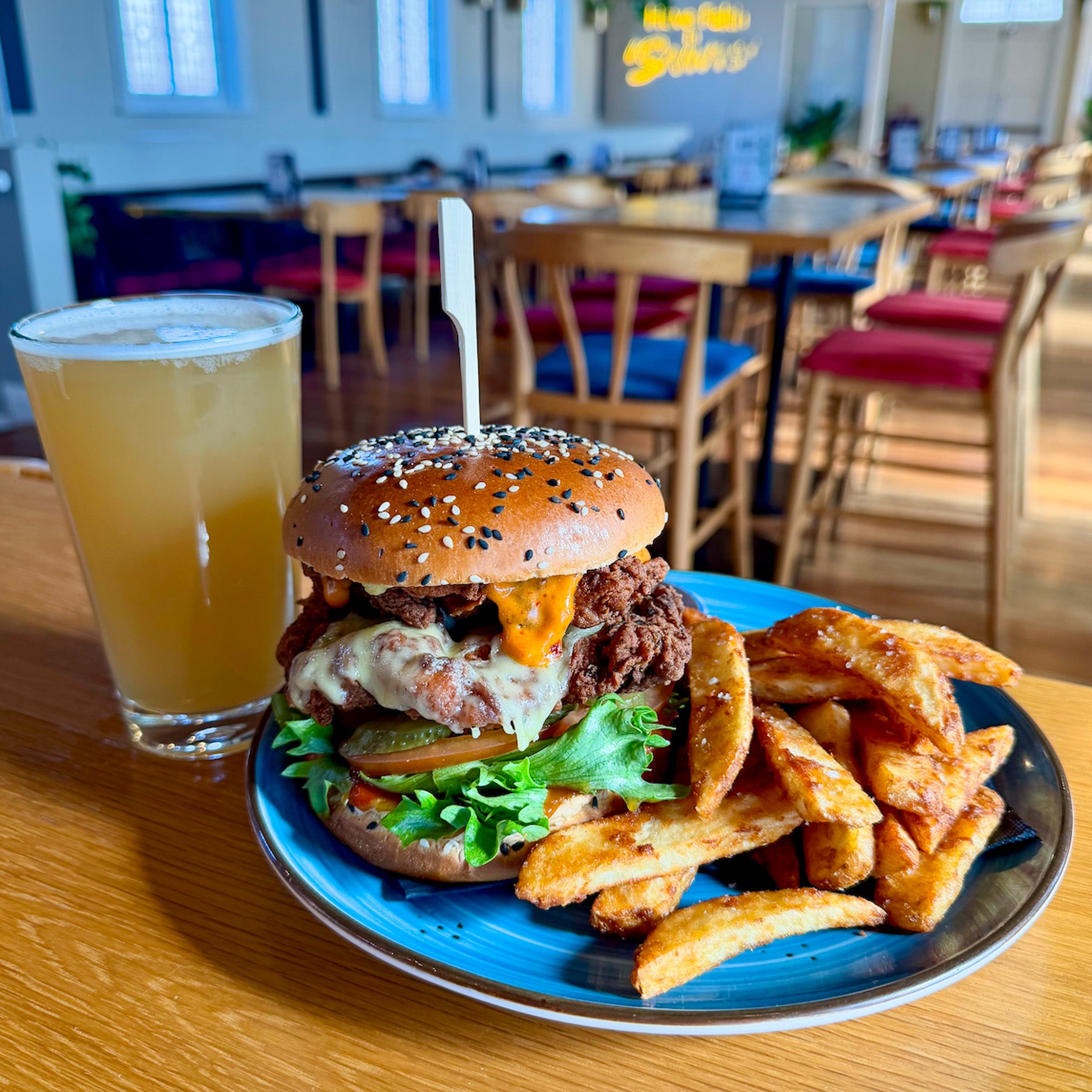 A loaded burger with cheese, lettuce, and pickles on a blue plate, served with thick fries and a glass of beer in a restaurant setting.