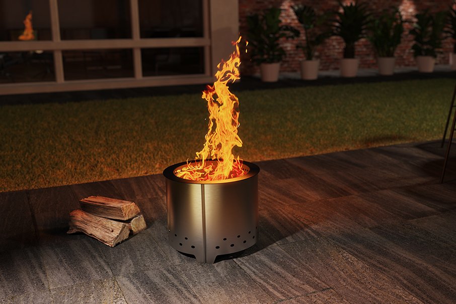 A cylindrical outdoor fire pit with a metallic finish, shown lit with bright orange flames on a dark patio. A small stack of wood logs sits to the left, with a grassy yard and potted plants in the background at night.