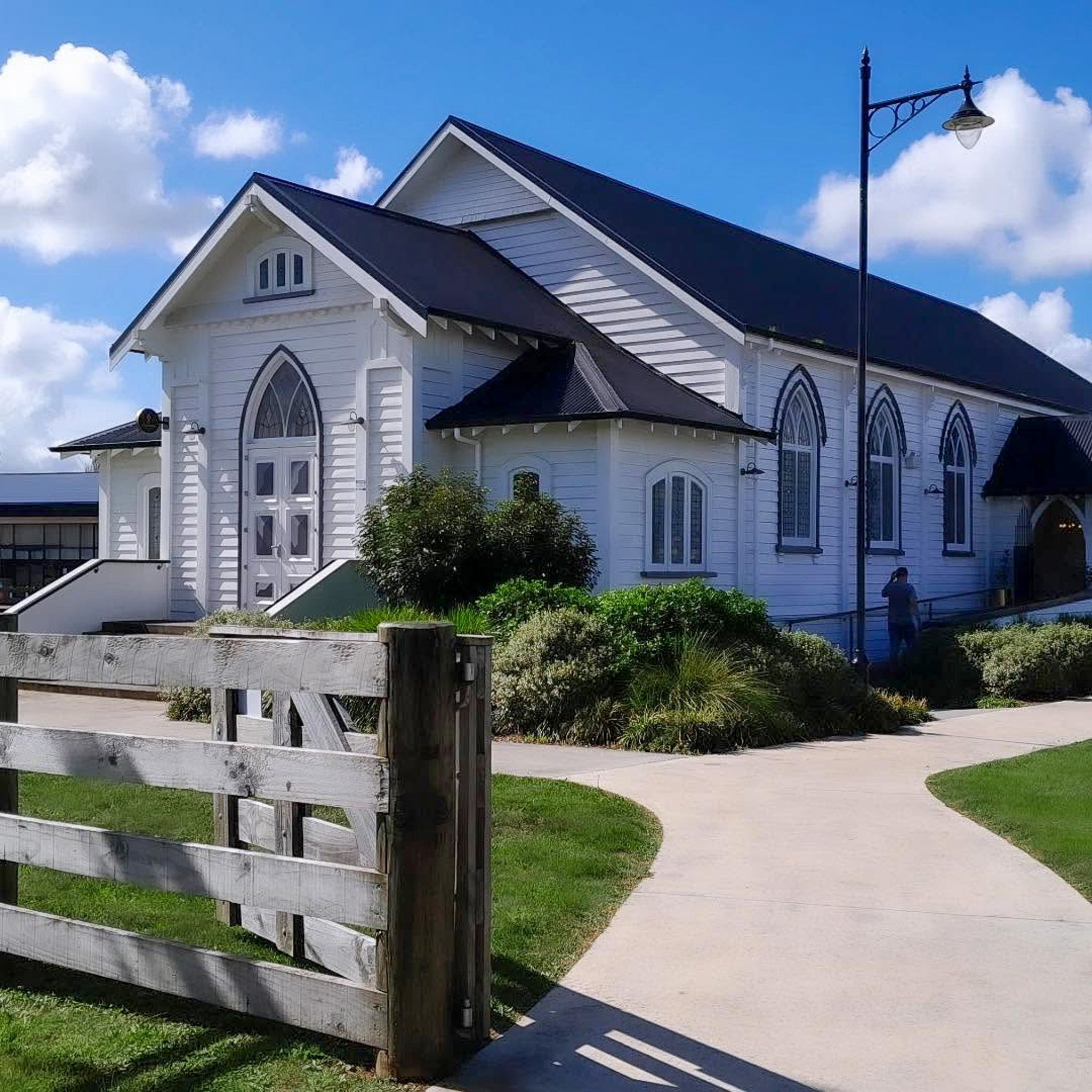 A white wooden church building with dark roofs, arched windows, a paved walkway, grass, shrubs, and a weathered wooden fence in the foreground under a blue sky with clouds.