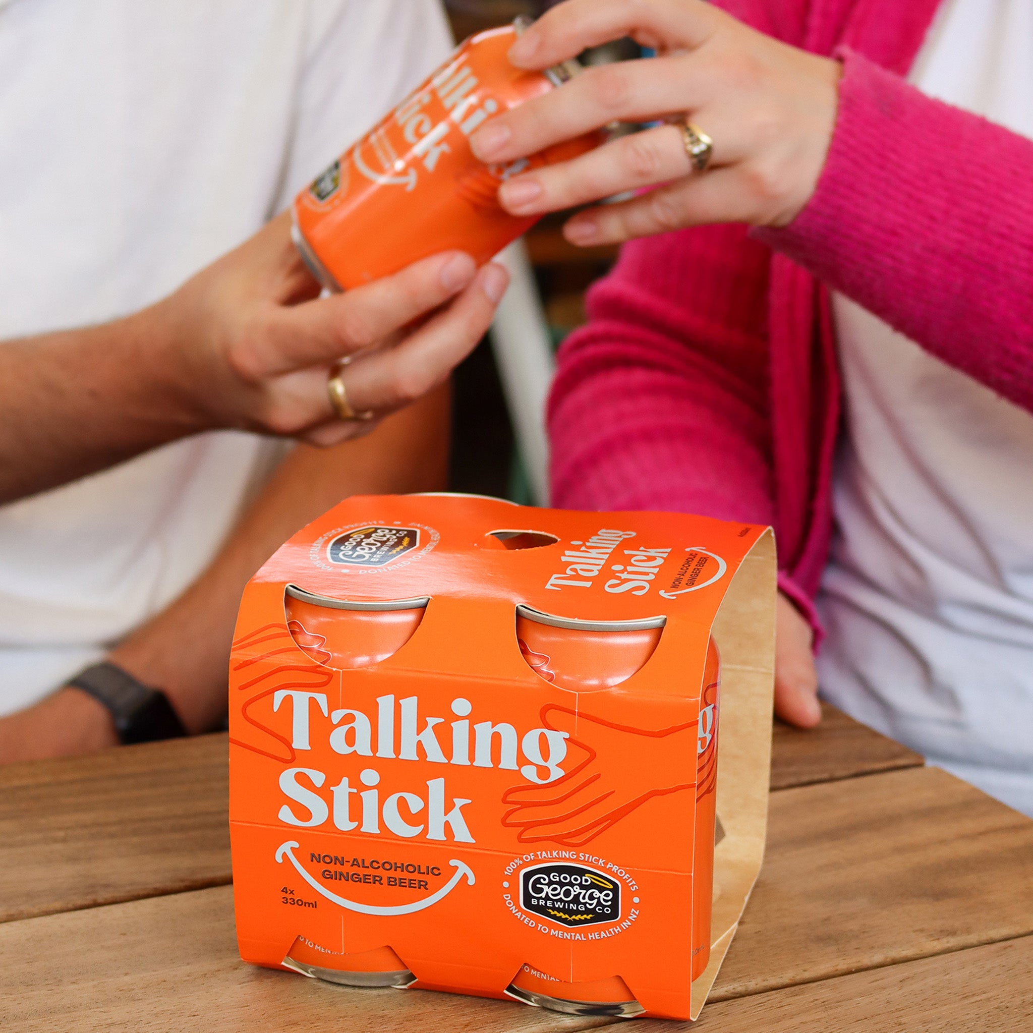 Two people holding 'Talking Stick' non-alcoholic ginger beer cans with a six-pack carton on a wooden table.