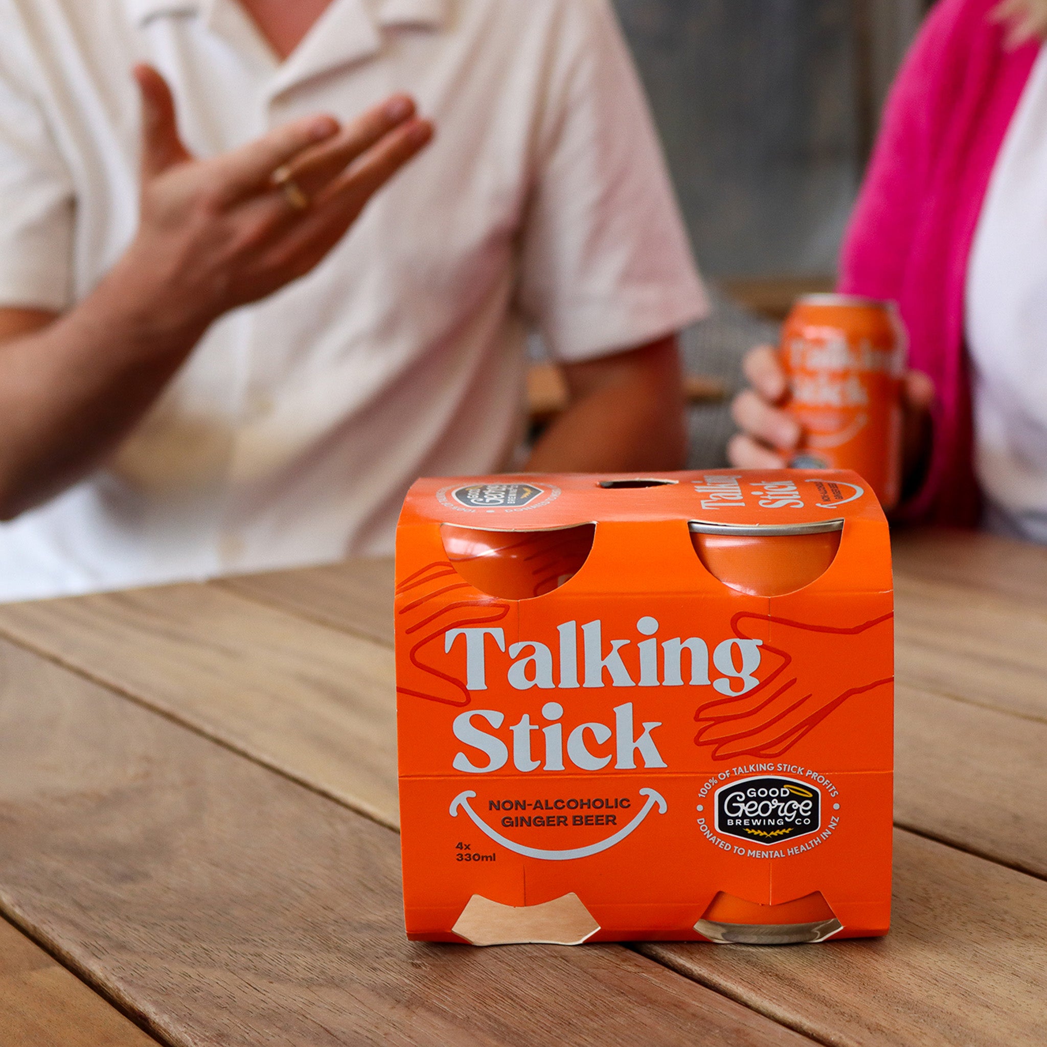Orange 'Talking Stick' non-alcoholic ginger beer packaging on a wooden table with people in the background.